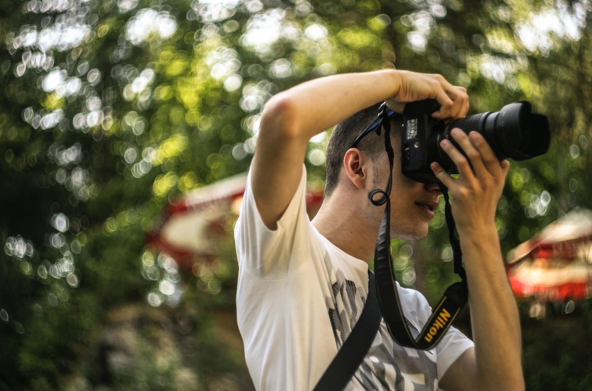 Młody mężczyzna w białej koszulce z aparatem Nikon w dłoniach, robiący zdjęcie na tle zieleni z efektem bokeh. Widoczny fragment czerwonego elementu w tle.
