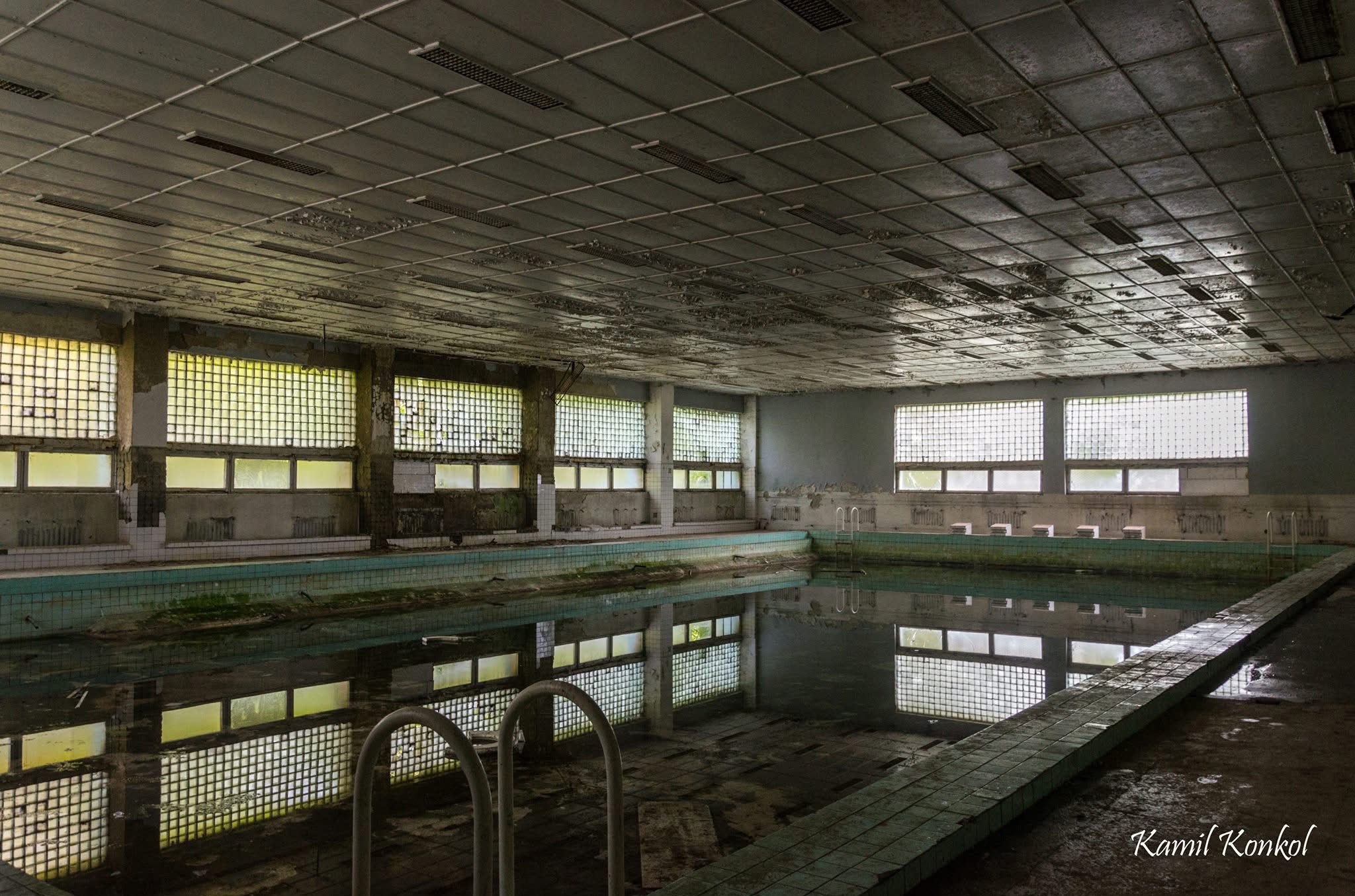 Opuszczony basen z widocznymi śladami zniszczeń, z odblaskami w wodzie i kratowanymi oknami. W tle widoczna sufitowa instalacja oświetleniowa.