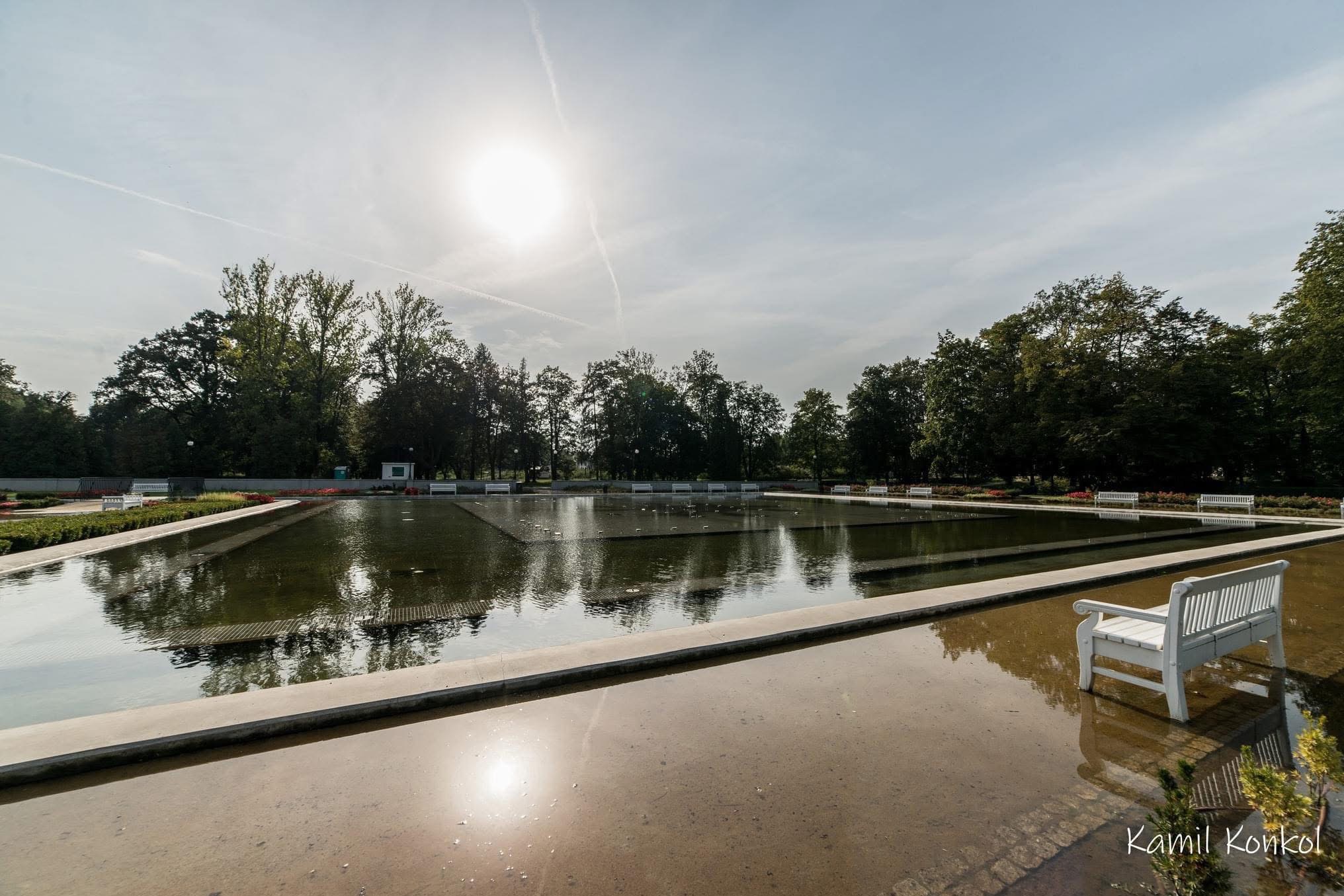 Parkowy zbiornik wodny z białymi ławkami w słoneczny dzień. Woda odbija niebo i drzewa. Jedna ławka stoi na pierwszym planie, częściowo zanurzona w wodzie.