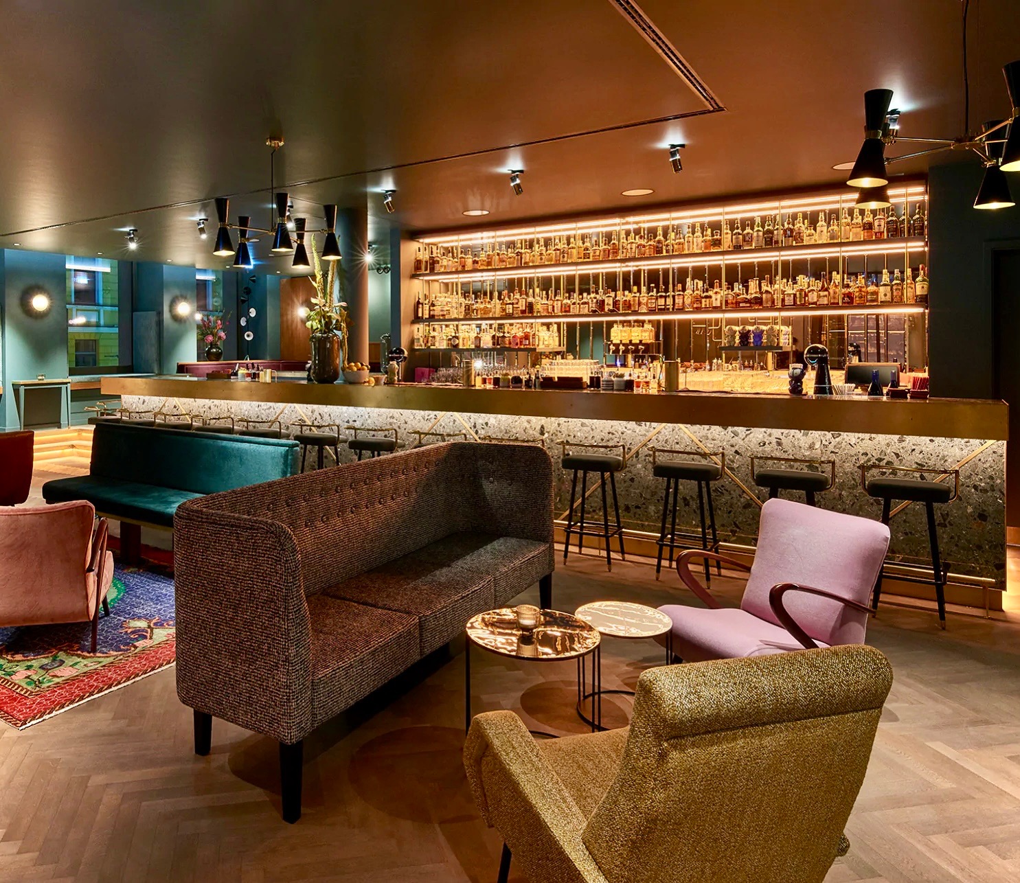 Eklektyczny bar z mozaikowym blatem, oświetlonymi półkami pełnymi butelek, pluszowymi sofami i fotelami w odcieniach różu i złota na parkiecie ułożonym we wzór jodełki.