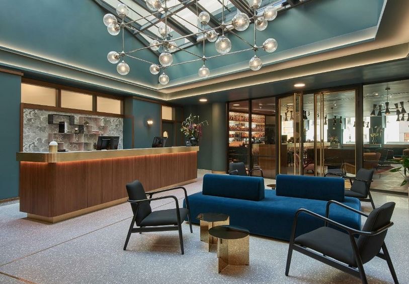 Recepcja hotelowa z drewnianą ladą, niebieską sofą i szklanym sufitem z designerską lampą. W tle widoczny bar za szklanymi drzwiami.