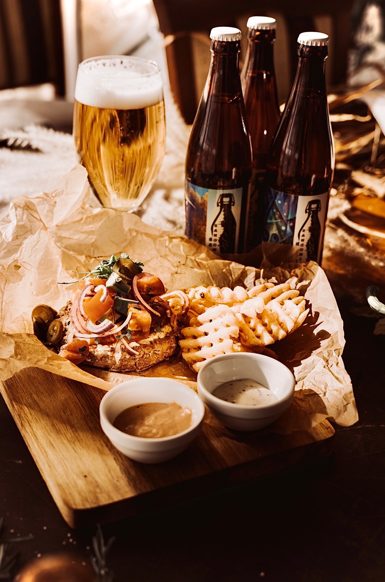 Apetyczna kompozycja: burger z warzywami, frytki waflowe, sosy i piwo kraftowe na drewnianej desce, w ciepłym oświetleniu. Gastronomiczna fotografia.