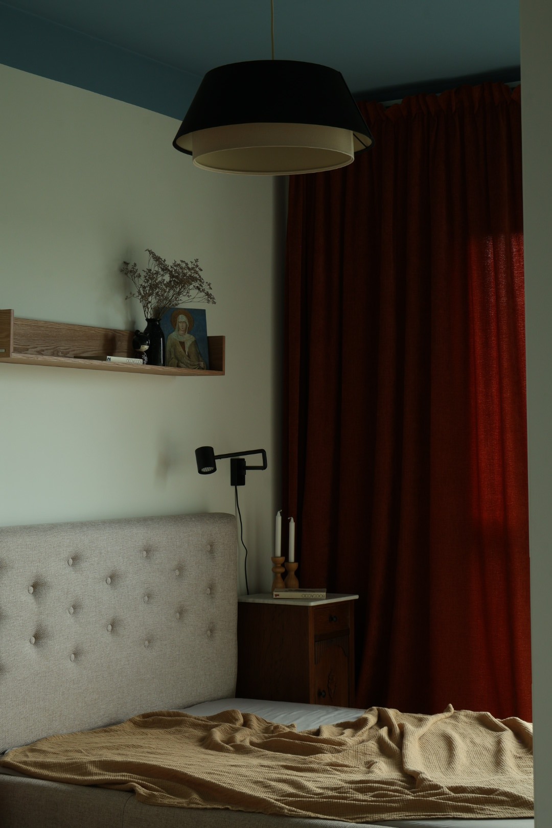 Neutralna sypialnia z beżowym zagłówkiem, drewnianą półką, czarną lampą i rdzawymi zasłonami. Minimalistyczny wystrój z akcentami vintage.