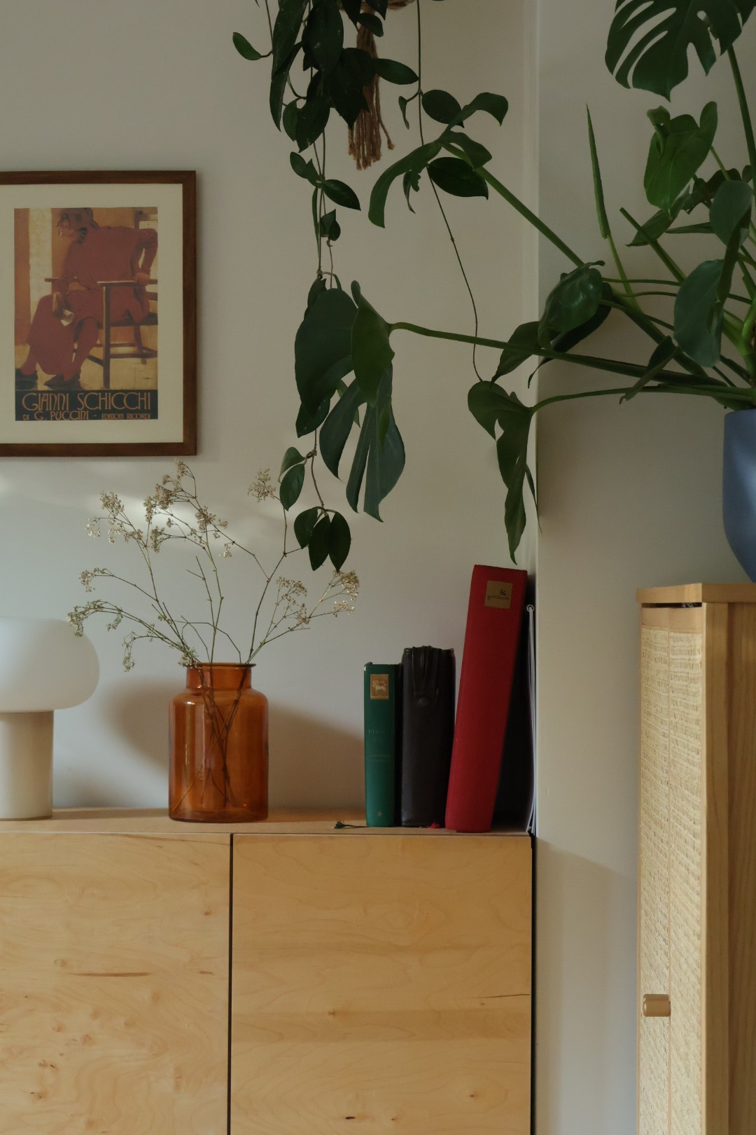 Jasny drewniany kredens z pomarańczowym wazonem i książkami. Nad nim wisząca roślina i plakat w ramie. Minimalistyczny wystrój wnętrza, ciepłe światło.