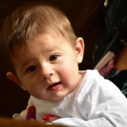 Portret niemowlęcia w białej koszulce, z zaciekawionym wyrazem twarzy, uchwycony w ciepłym oświetleniu, z delikatnym rozmyciem tła. Dziecko trzyma zabawkę.