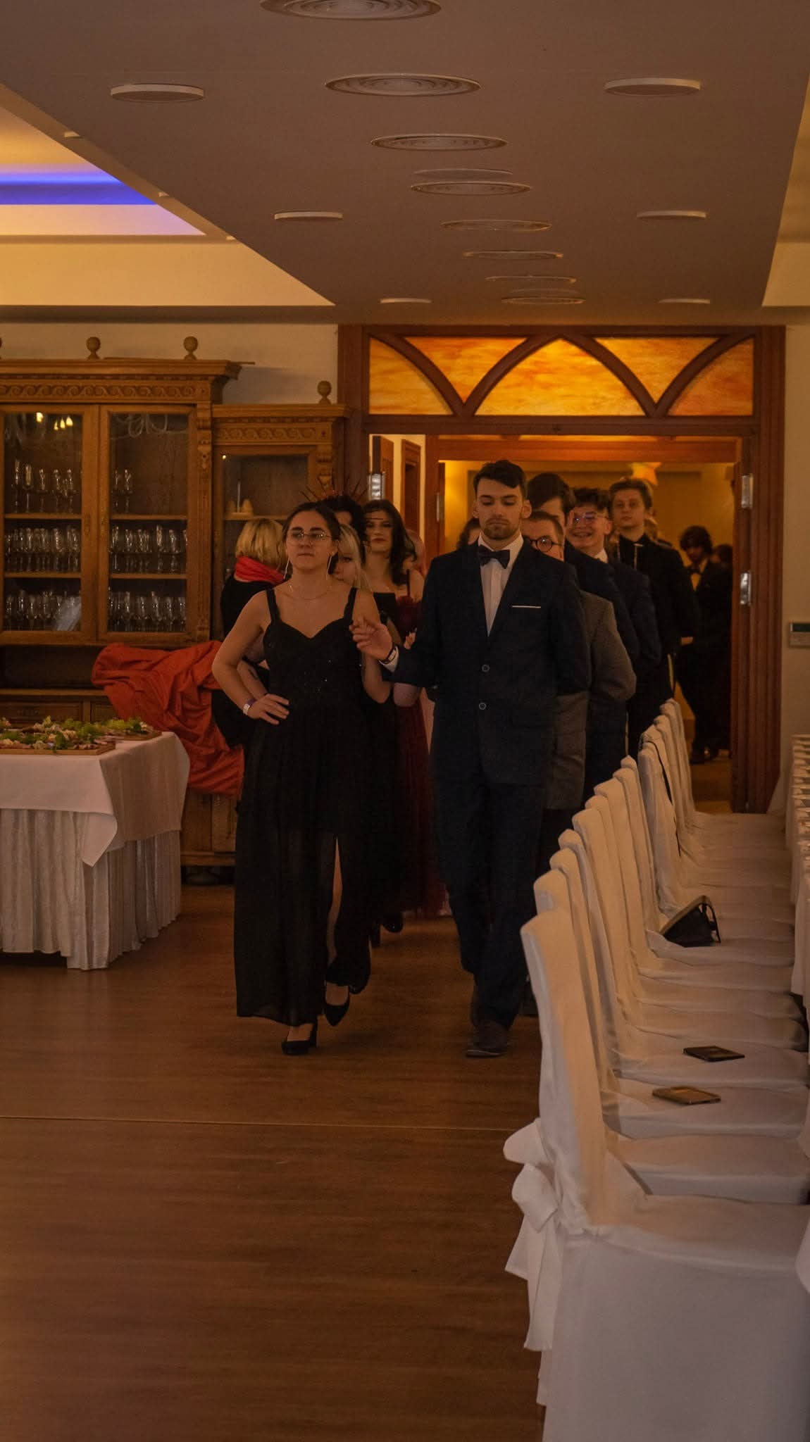 Uczestnicy studniówki w eleganckich strojach wchodzą na salę balową, widoczne nakryte stoły i dekoracje. Ujęcie z perspektywy gości.