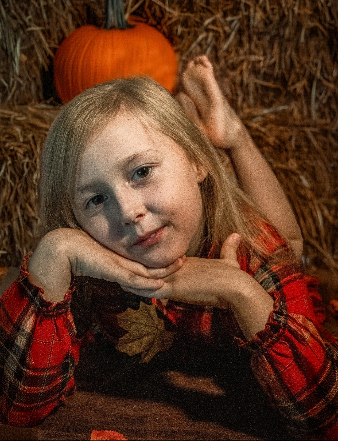 Portret dziewczynki w czerwonej kraciastej sukience, leżącej na sianie z dynią w tle. Jesienna sesja w ciepłych barwach, z liściem klonu na sukience.