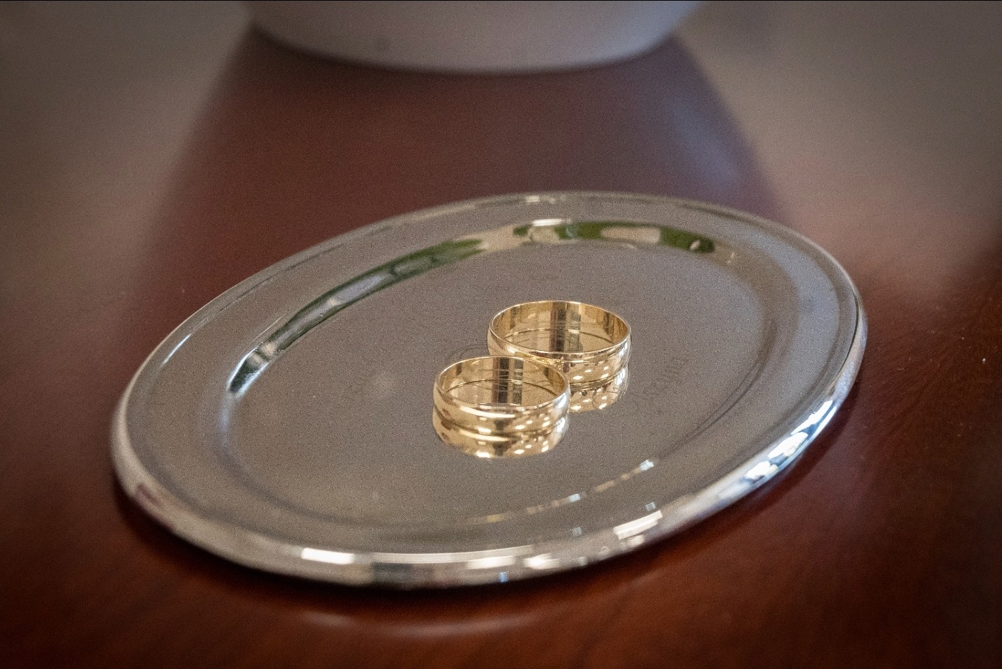Dwie złote obrączki ślubne leżą na srebrnej, owalnej tacy na tle ciemnego blatu. Obrączki są ustawione blisko siebie, a ich faktura jest delikatnie widoczna.