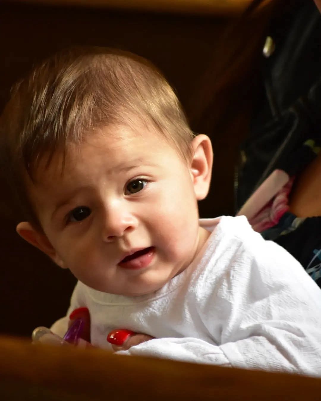 Portret niemowlęcia w białej koszulce, z zaciekawionym wyrazem twarzy, uchwycony w ciepłym oświetleniu, z delikatnym rozmyciem tła. Dziecko trzyma zabawkę.