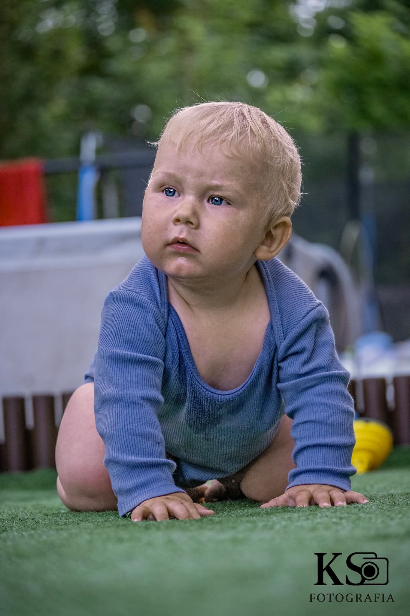 Niemowlę z blond włosami i niebieskimi oczami w niebieskim ubranku raczkuje na zielonej trawie, patrząc w górę. W tle rozmyte otoczenie.