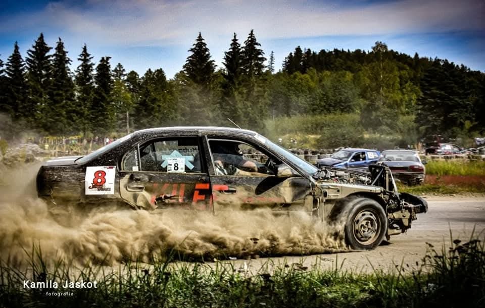 Rozmyty obraz zniszczonego samochodu rajdowego nr 8 w akcji, unoszącego tumany kurzu na tle zieleni i innych pojazdów. Ujęcie dynamiczne.