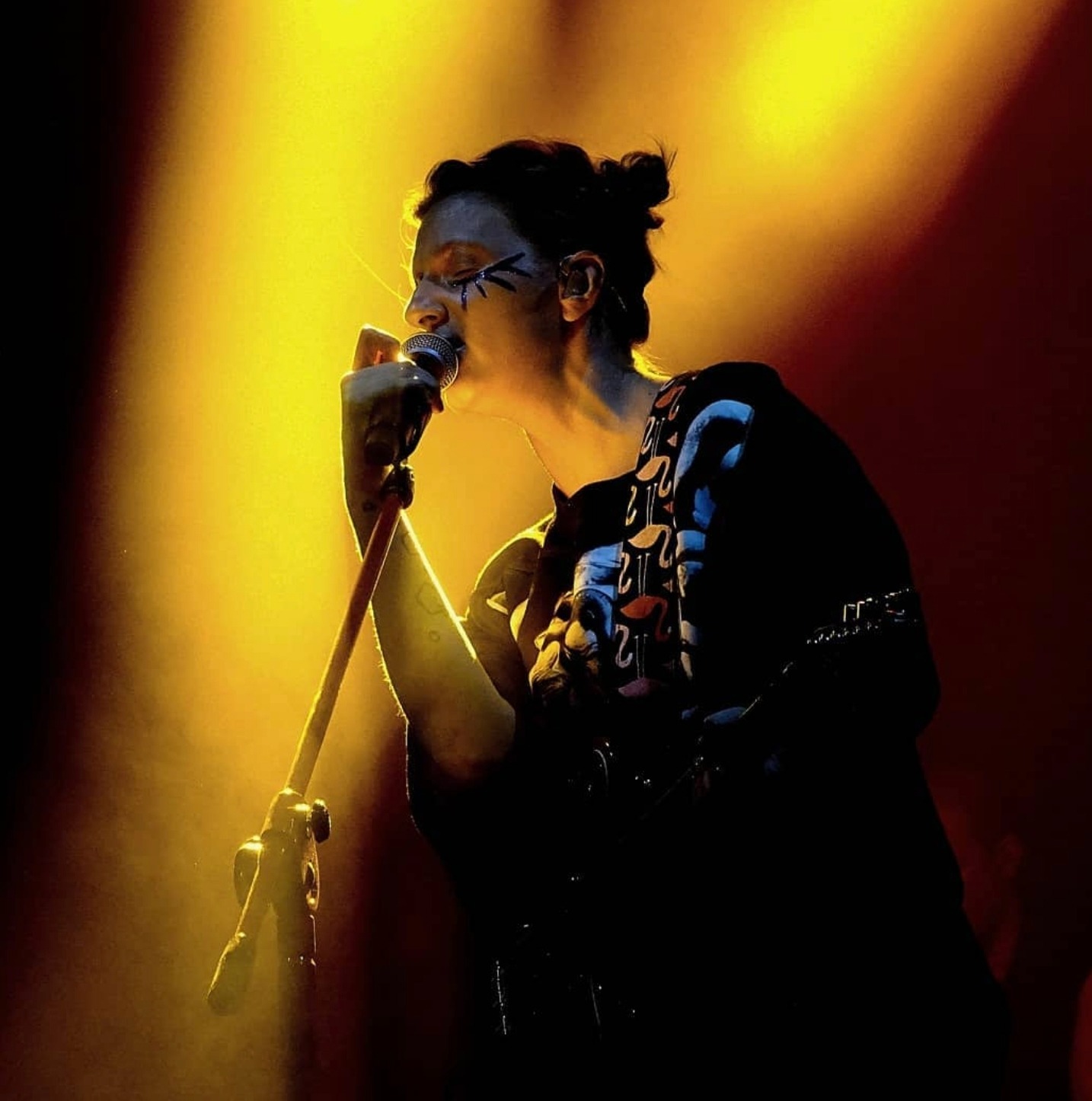 Piosenkarka z mocnym makijażem scenicznym śpiewa do mikrofonu w intensywnym, żółtym świetle reflektorów podczas koncertu.