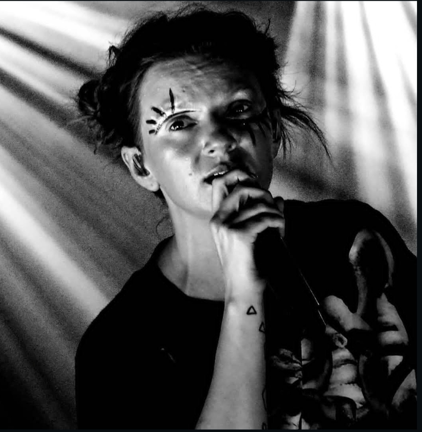 Czarno-białe zdjęcie wokalistki z artystycznym makijażem, trzymającej mikrofon na scenie w blasku reflektorów. Widoczny tatuaż na ręce.