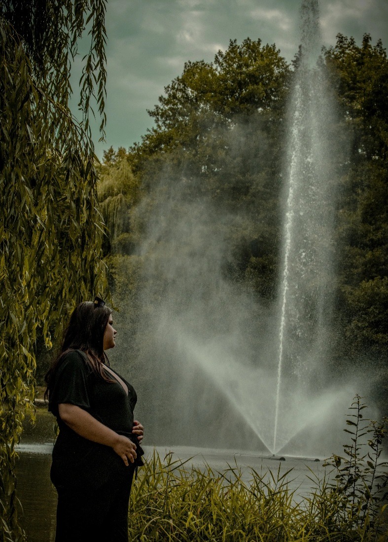 Kobieta w czarnej sukience pozuje na tle fontanny w parku, z profilu. Ujęcie z dołu, z widocznymi trawami i drzewami.