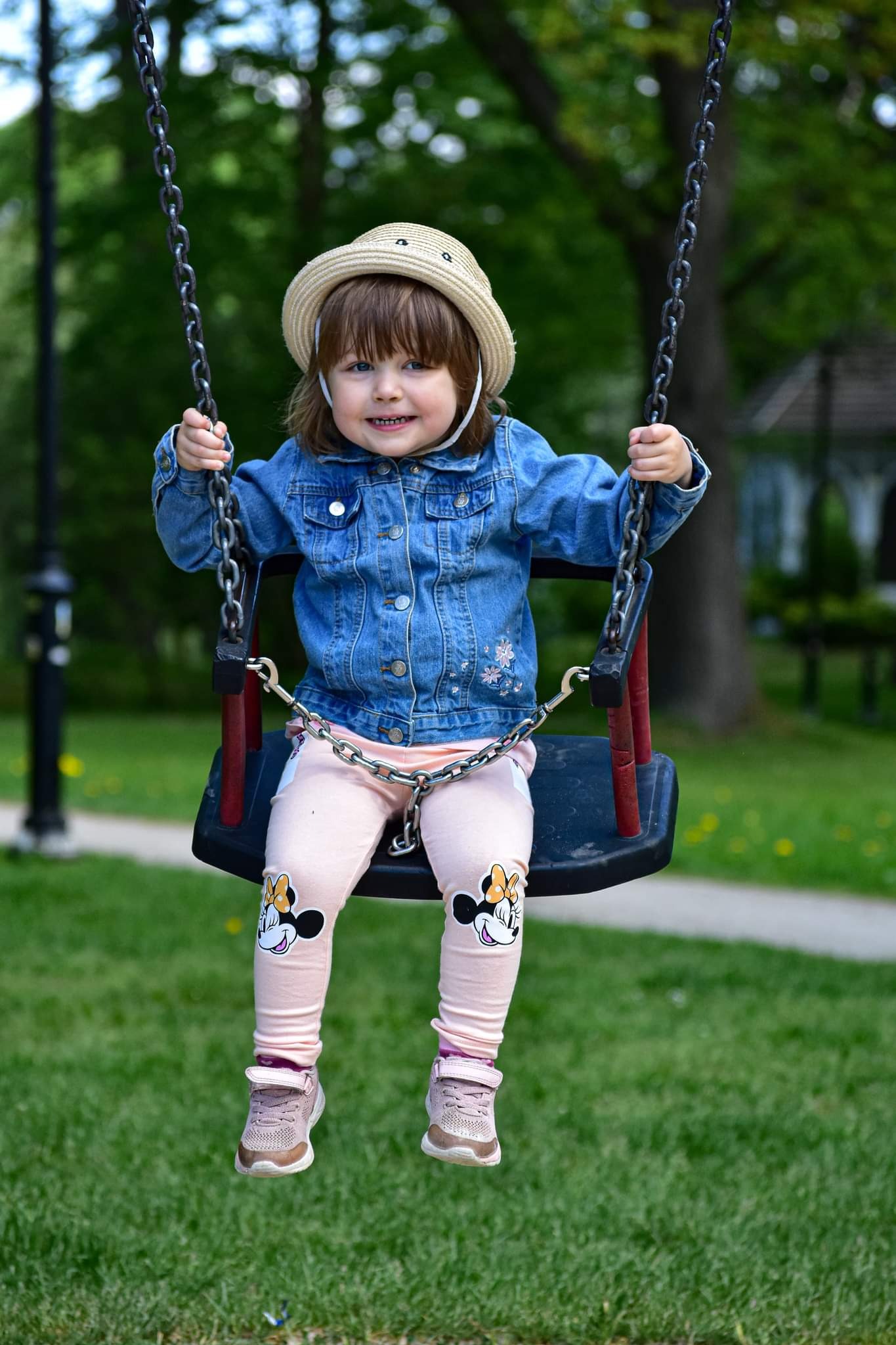 Uśmiechnięta dziewczynka w dżinsowej kurtce i kapeluszu siedzi na huśtawce na tle zieleni. Na spodniach ma aplikacje Myszki Minnie.