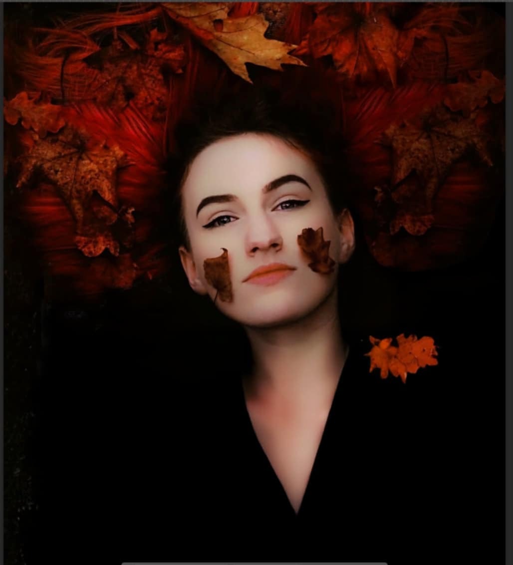 Portret kobiety z liśćmi na twarzy i we włosach, w ciemnej tonacji. Artystyczna sesja w jesiennej scenerii, z mocnym makijażem oczu i wyrazistym oświetleniem.
