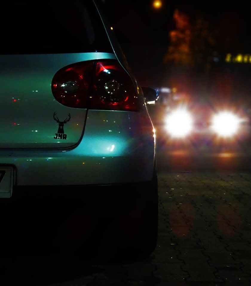 Tył srebrnego samochodu z naklejką jelenia JNR w nocnej scenerii, światła innego pojazdu w tle, oświetlenie uliczne.