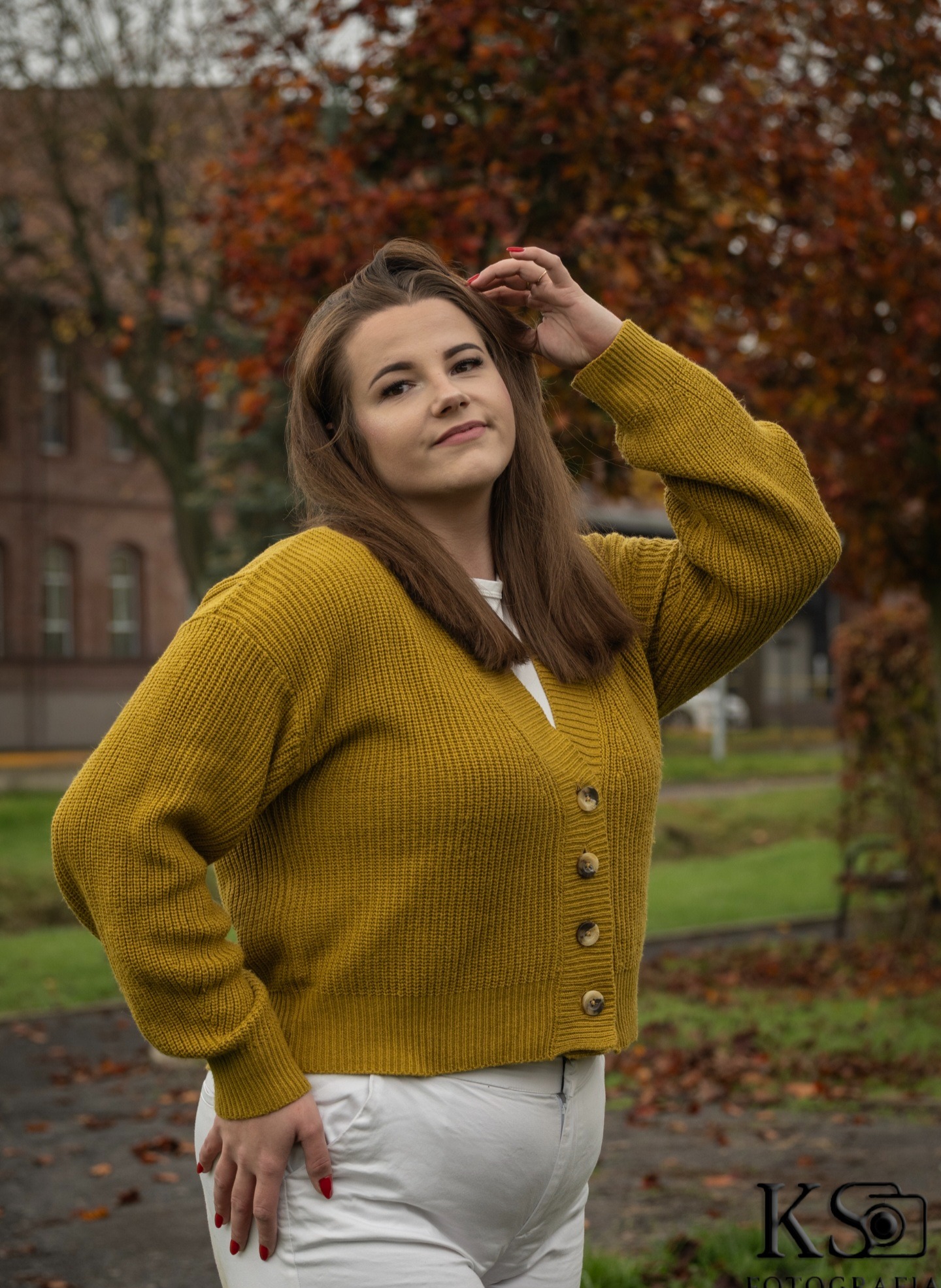 Portret kobiety w musztardowym kardiganie na tle jesiennego krajobrazu. Uśmiechnięta, z ręką we włosach, pozuje w plenerze. Widoczny budynek w tle i logo fotografa.