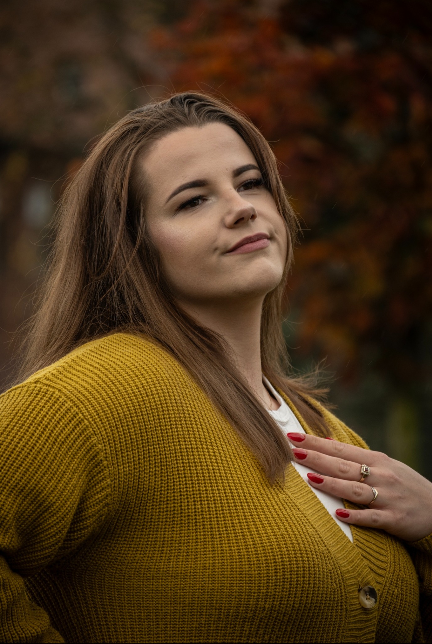 Portret kobiety w musztardowym swetrze na tle jesiennych drzew. Uwagę przyciągają czerwone paznokcie i pierścionki na dłoni. Delikatny uśmiech i naturalny makijaż.