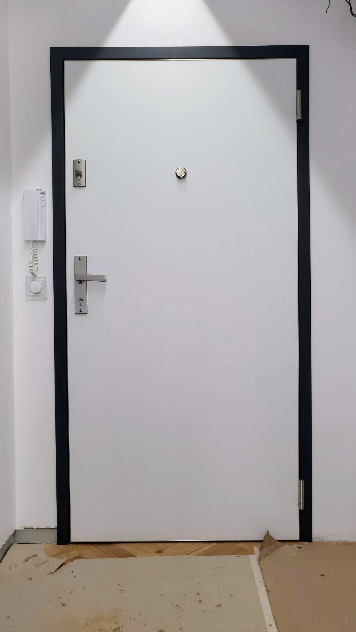 Zamknięte, białe drzwi wejściowe z czarną ościeżnicą, widoczne od frontu. Na ścianie domofon. Podłoga zabezpieczona kartonem. Widoczne zawiasy i klamka.