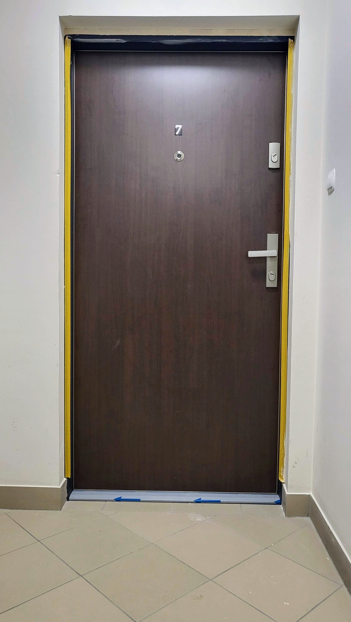 Ciemne, drewnopodobne drzwi z numerem '7' i srebrną klamką, zabezpieczone żółtą taśmą malarską. Widoczna podłoga z płytek i fragment białej ściany.