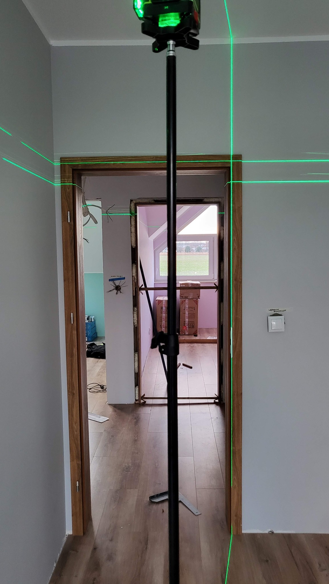Laser krzyżowy wyznacza pion i poziom na ścianie podczas montażu ościeżnicy drewnianej w nowym wnętrzu z panelami. Widoczne narzędzie pomiarowe.