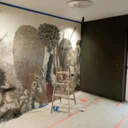 SEBA SERWIS DOMOWY Firma Remontowo-Budowlana Tomasik Sebastian - Pokój w trakcie malowania z motywem roślinnym na ścianie. Widoczna drabina, taśmy malarskie i częściowo pomalowana ściana na ciemny kolor. Prace wykończeniowe.