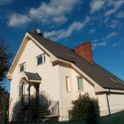 SEBA SERWIS DOMOWY Firma Remontowo-Budowlana Tomasik Sebastian - Jasny dom jednorodzinny z ciemną dachówką i ceglanym kominem na tle błękitnego nieba z chmurami. Widoczne schody wejściowe z metalową barierką.
