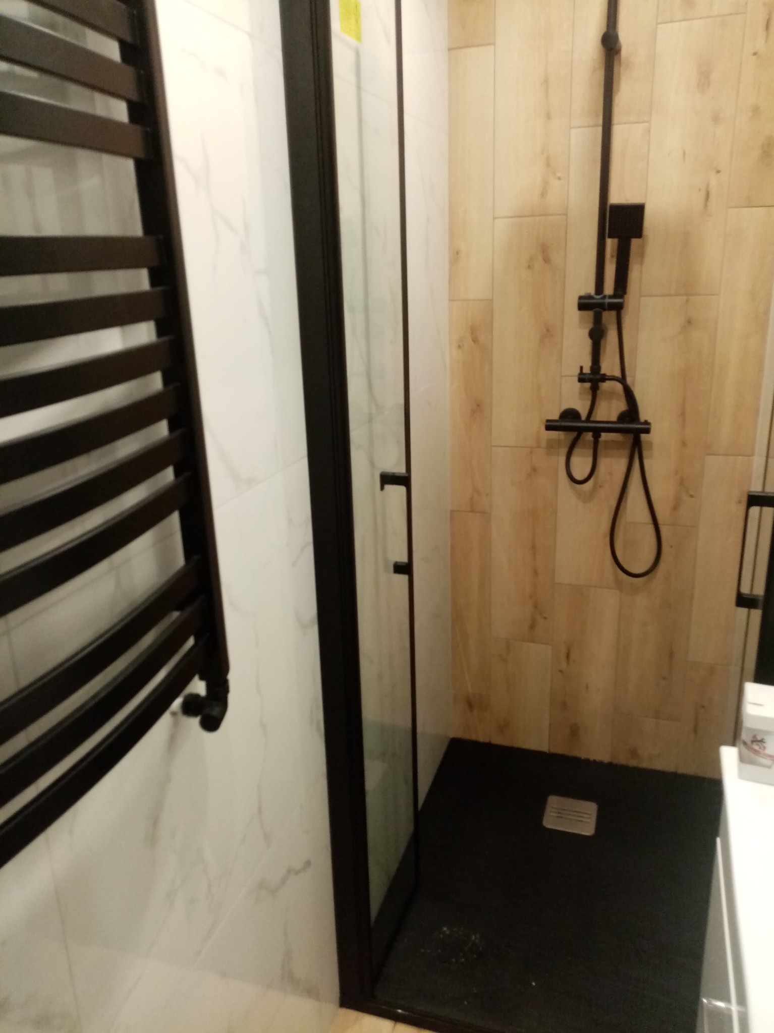 Nowoczesna kabina prysznicowa z czarną armaturą i grzejnikiem drabinkowym. Białe marmurowe płytki kontrastują z drewnopodobnymi panelami.