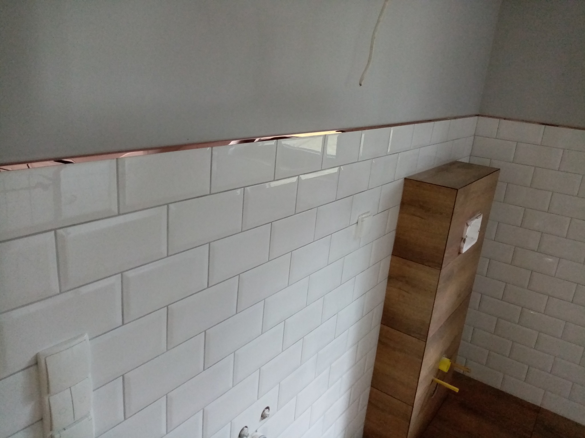 Białe, prostokątne płytki ułożone w łazience w Wytomyślu, z dekoracyjną listwą w kolorze miedzi. Widoczna zabudowa z drewnopodobnych płytek.