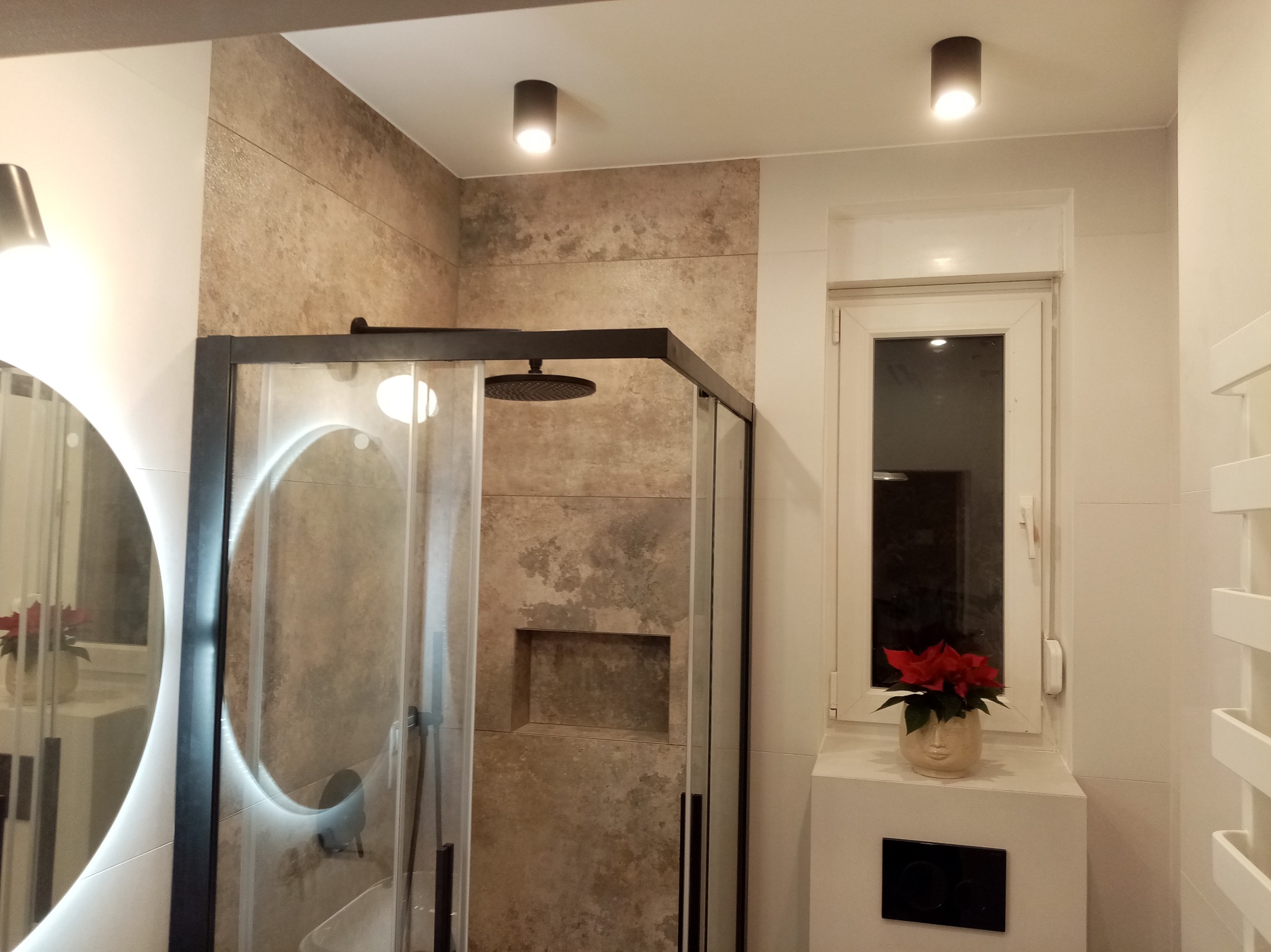 Nowoczesna łazienka z kabiną typu walk-in, beżowymi płytkami imitującymi kamień, oświetleniem punktowym i okrągłym lustrem z podświetleniem LED.