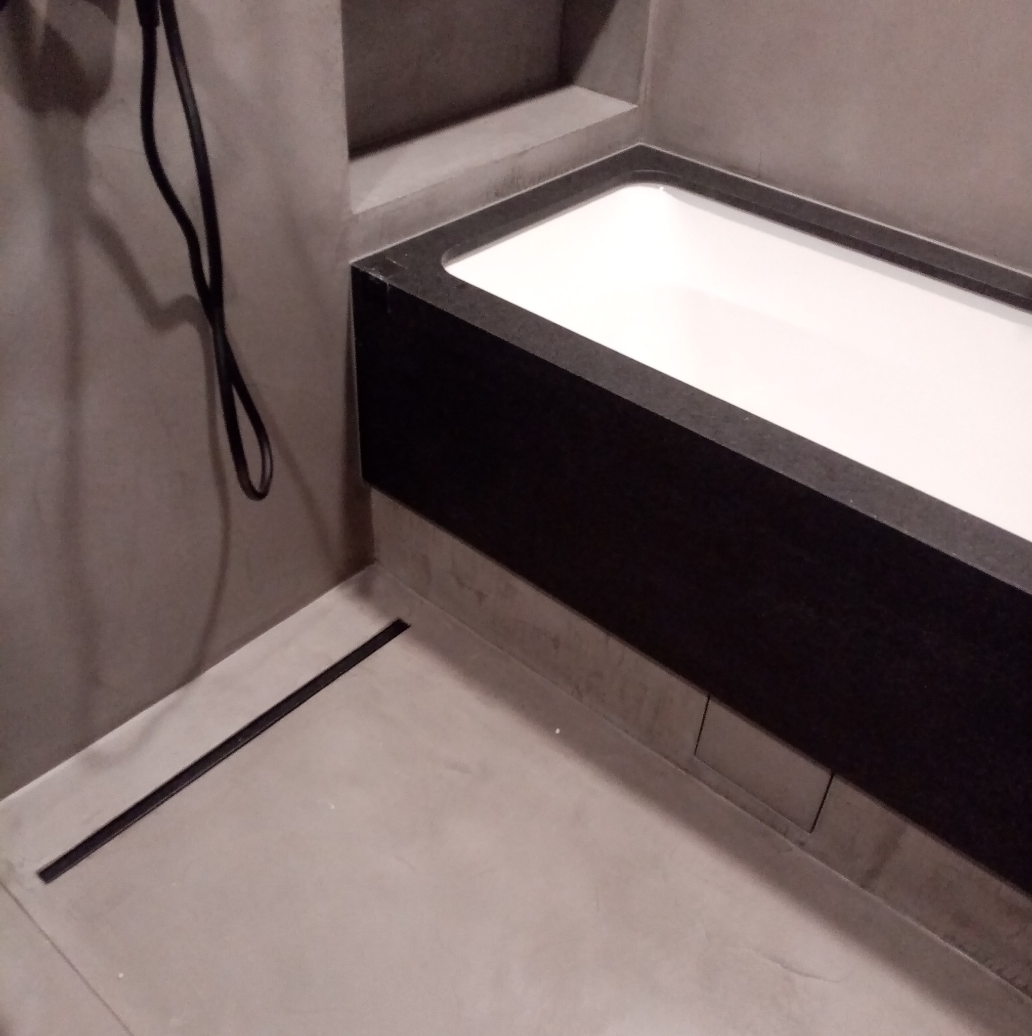 Minimalistyczna łazienka z czarną obudową wanny i odpływem liniowym w podłodze, utrzymana w szarościach. Widoczny fragment słuchawki prysznicowej.