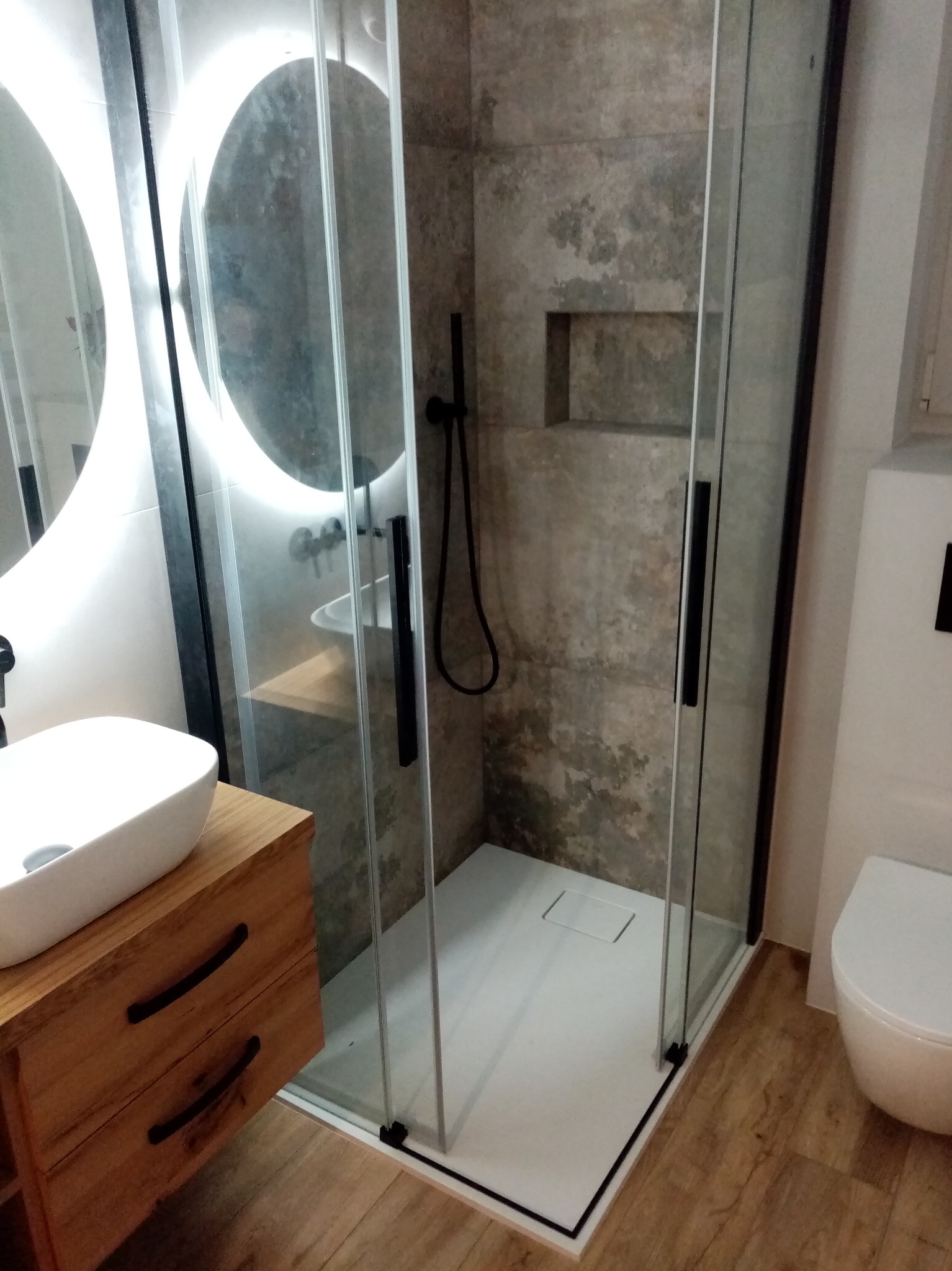 Nowoczesna łazienka z kabiną prysznicową typu walk-in, umywalką nablatową na drewnianej szafce i okrągłym lustrem z podświetleniem LED.