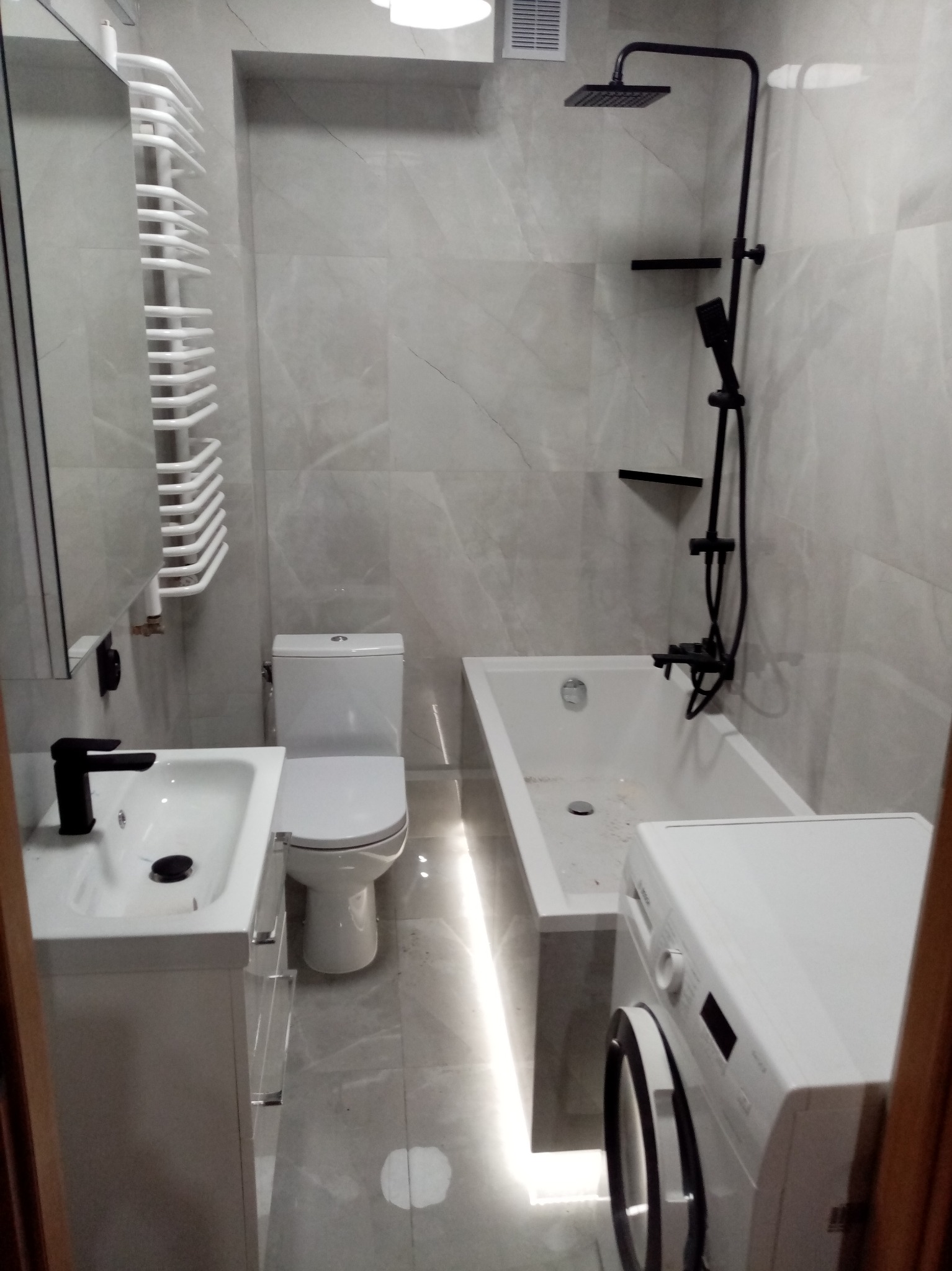 Nowoczesna łazienka po remoncie: szare płytki, biała armatura, czarna bateria i deszczownica. Zwraca uwagę grzejnik drabinkowy i oświetlenie LED pod wanną.