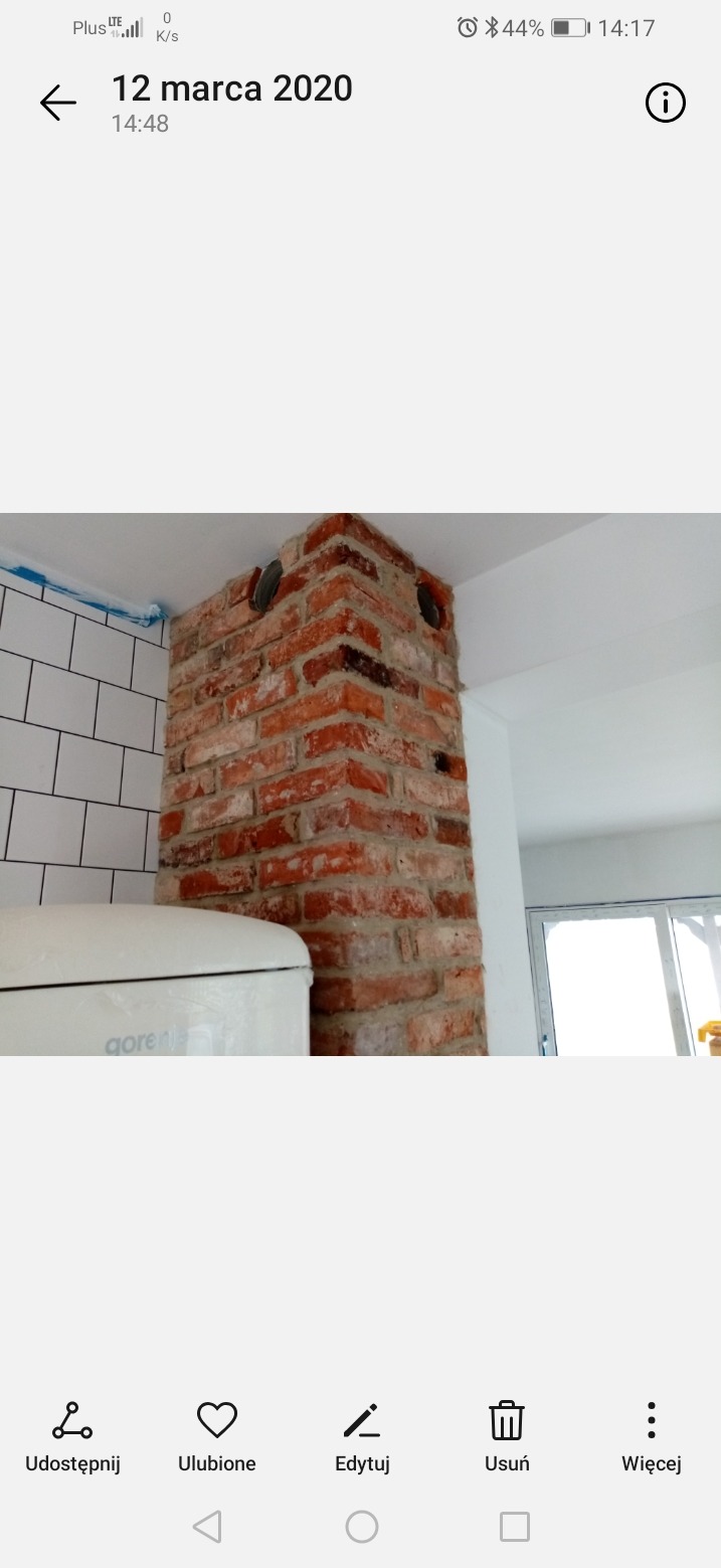 Wyeksponowany fragment ceglanej ściany z widocznymi otworami instalacyjnymi, sąsiadujący z białą glazurą i sprzętem AGD. Element wykończenia wnętrza w stylu industrialnym.