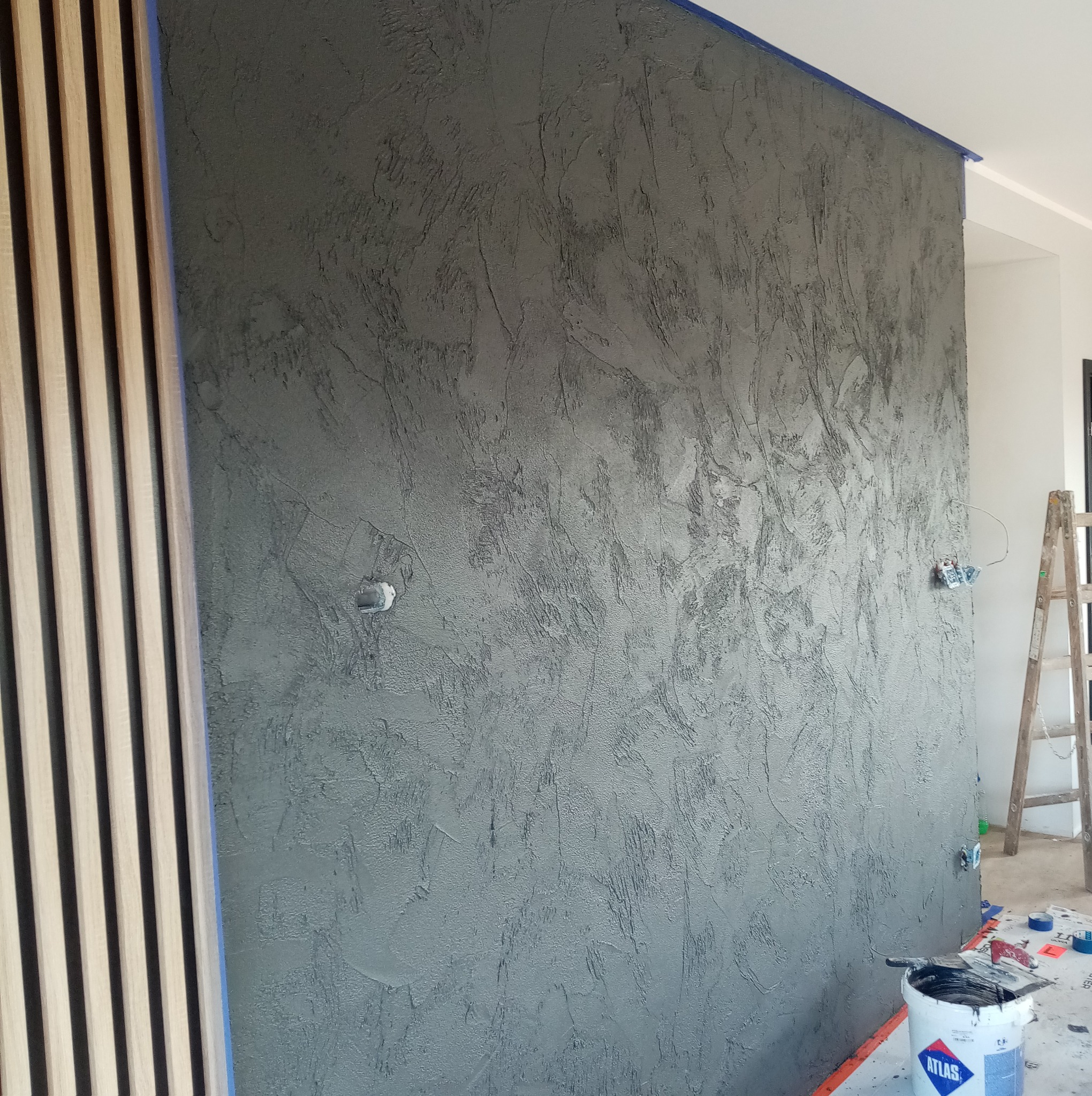 Ściana po szpachlowaniu dekoracyjnym w odcieniach szarości, z widoczną strukturą i elementami wykończeniowymi, takimi jak listwy drewniane i zabezpieczone gniazdka.