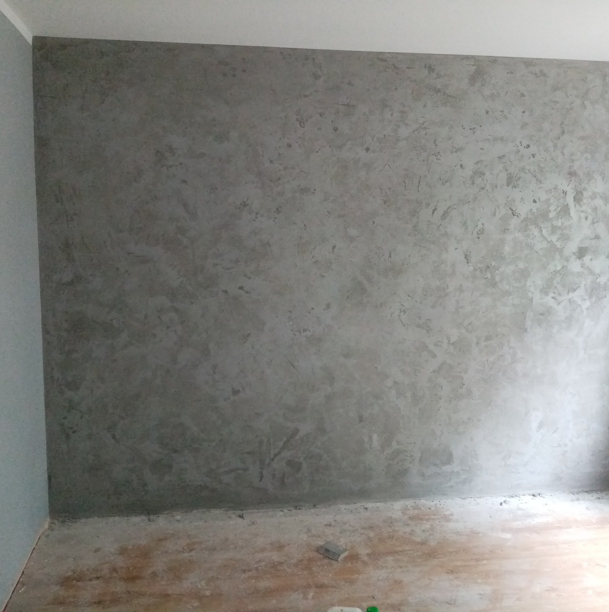 Ściana z efektem dekoracyjnym w odcieniach szarości, widoczne niedoskonałości podłogi, sufit biały. Styl loftowy, surowy wygląd.