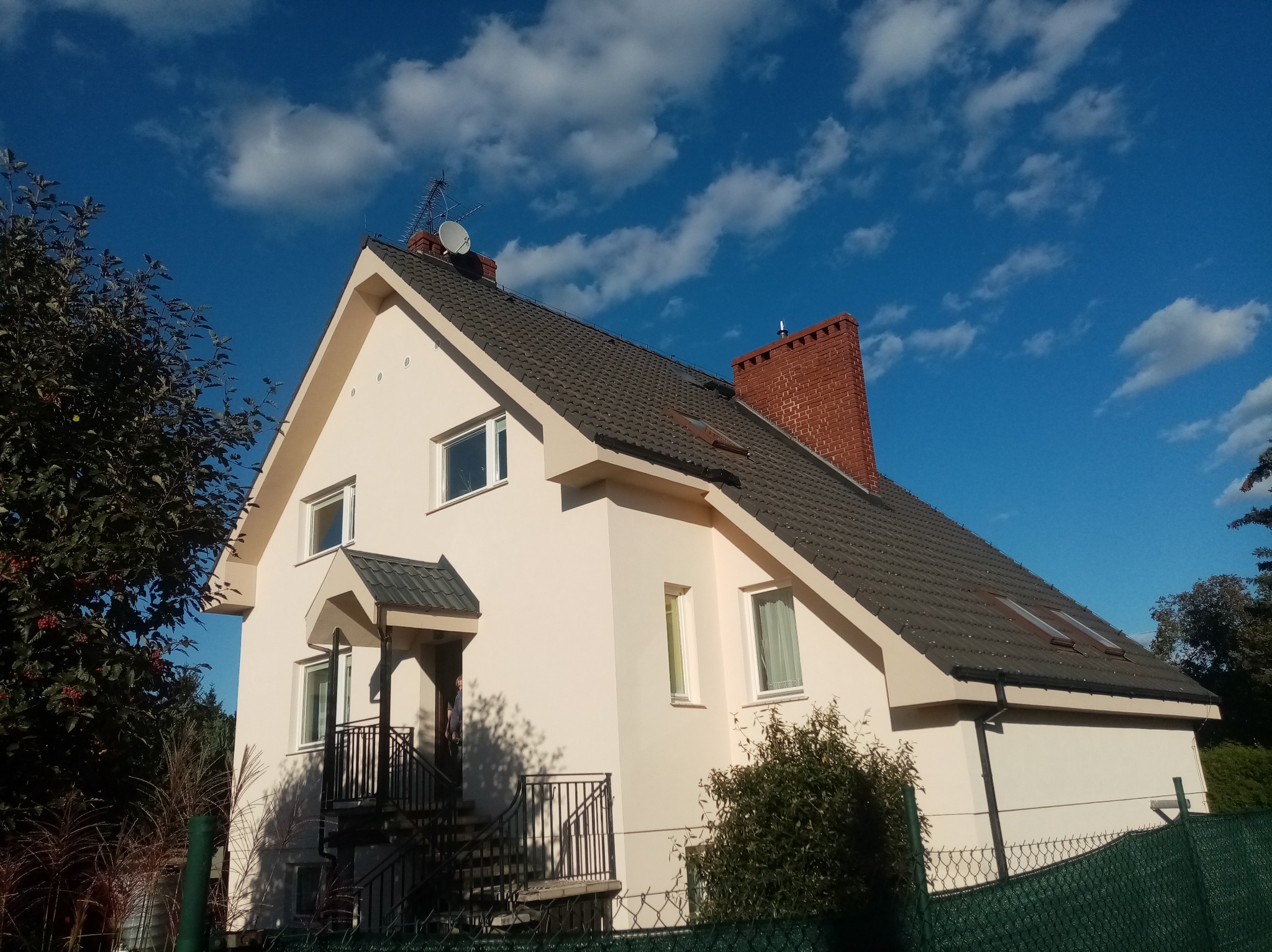 Jasny dom jednorodzinny z ciemną dachówką i ceglanym kominem na tle błękitnego nieba z chmurami. Widoczne schody wejściowe z metalową barierką.