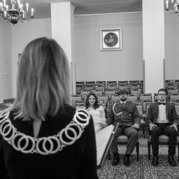 LESZEK ŚWIĘCH TWÓJ FOTOGRAFIK - Czarno-białe zdjęcie z ceremonii ślubnej: urzędniczka z łańcuchem, para młoda i świadek siedzą na krzesłach w sali z żyrandolami i obrazem na ścianie.