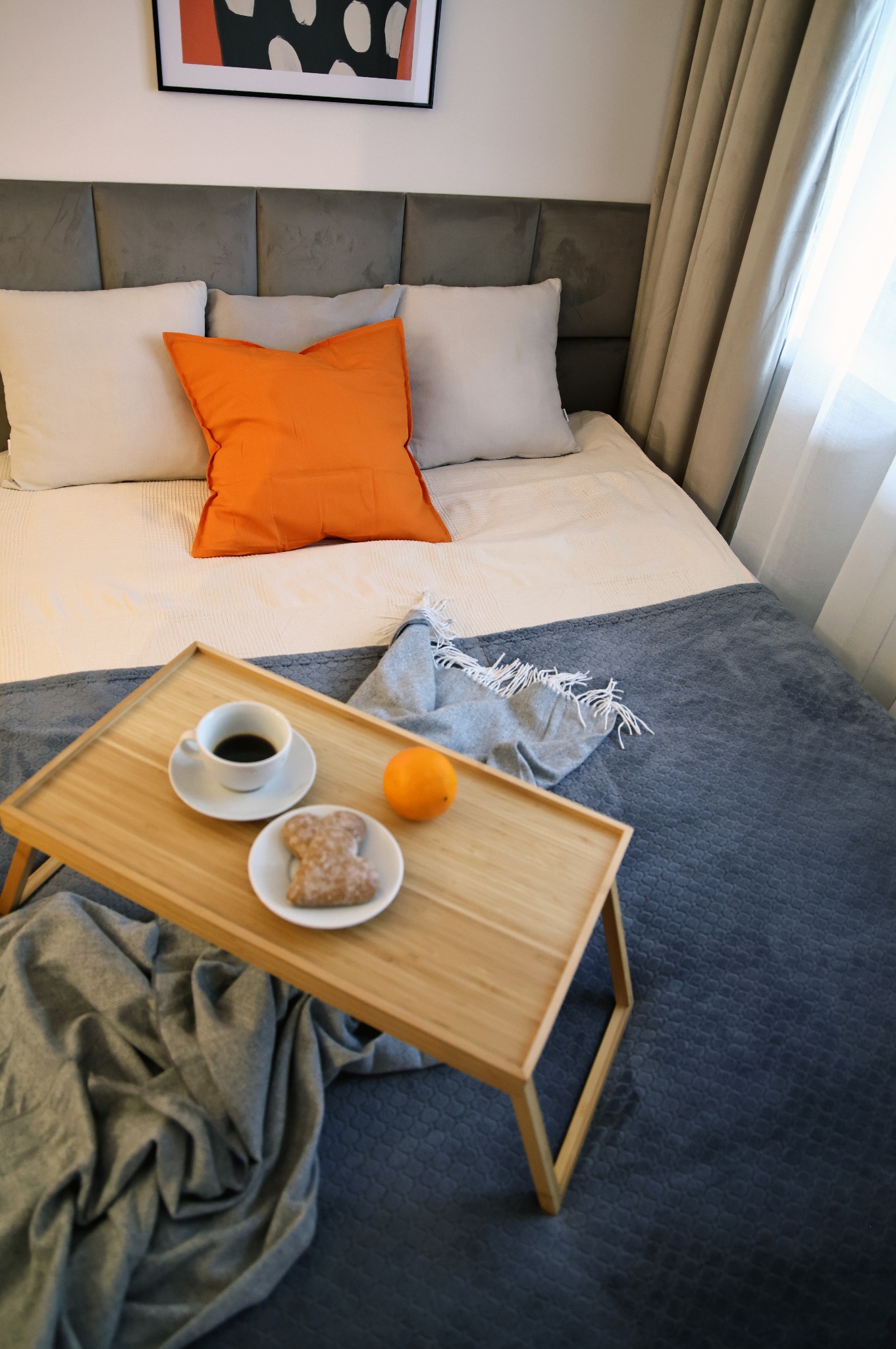 Jasne wnętrze sypialni z łóżkiem, na którym znajduje się drewniana taca z kawą, ciastkiem i pomarańczą. W tle szare wezgłowie i zasłony.