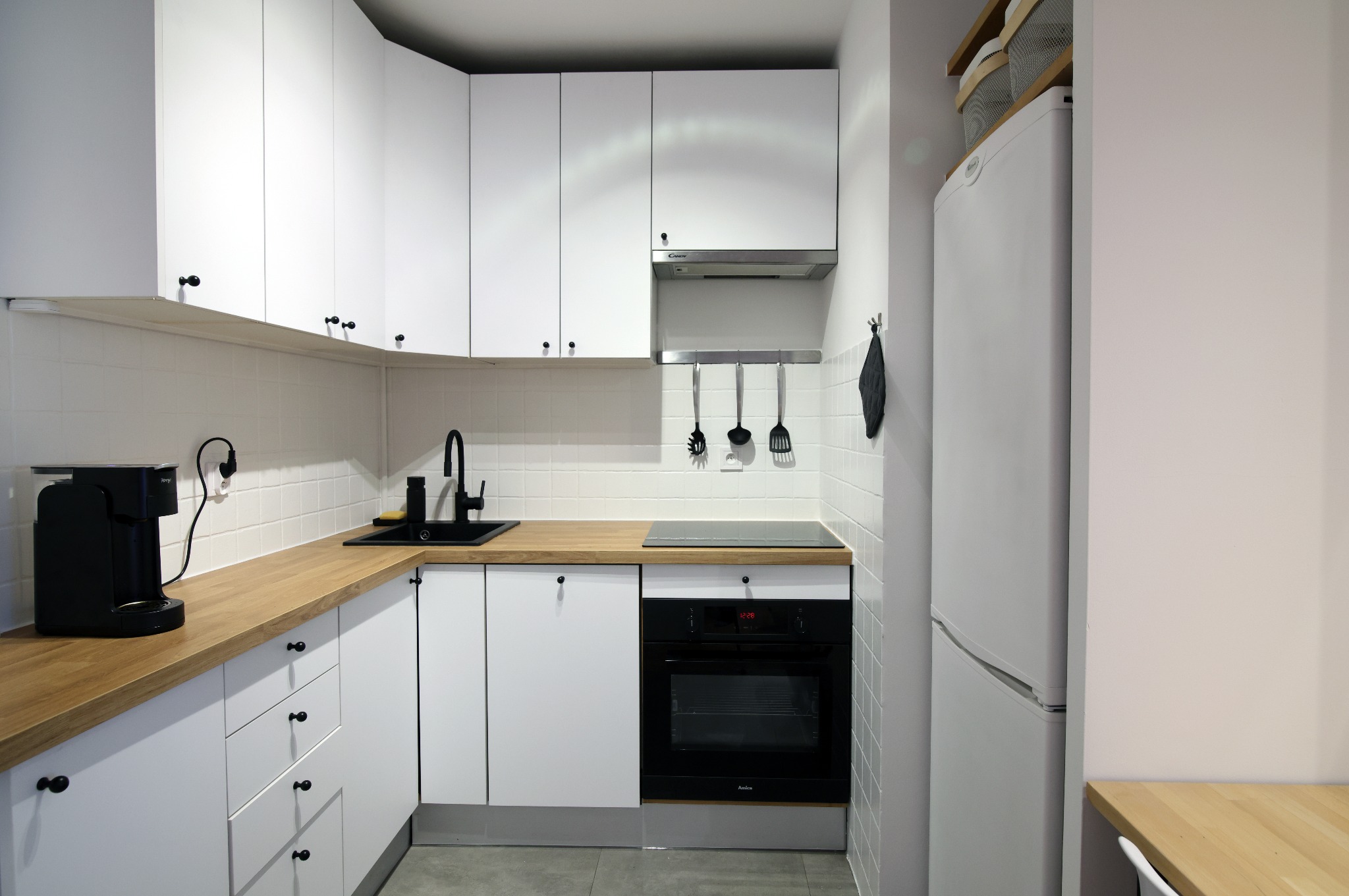 Nowoczesna, biała kuchnia z drewnianym blatem, czarną armaturą i sprzętami AGD. Minimalistyczny design, płytki na ścianie i szare płytki na podłodze. Widok na całe pomieszczenie.