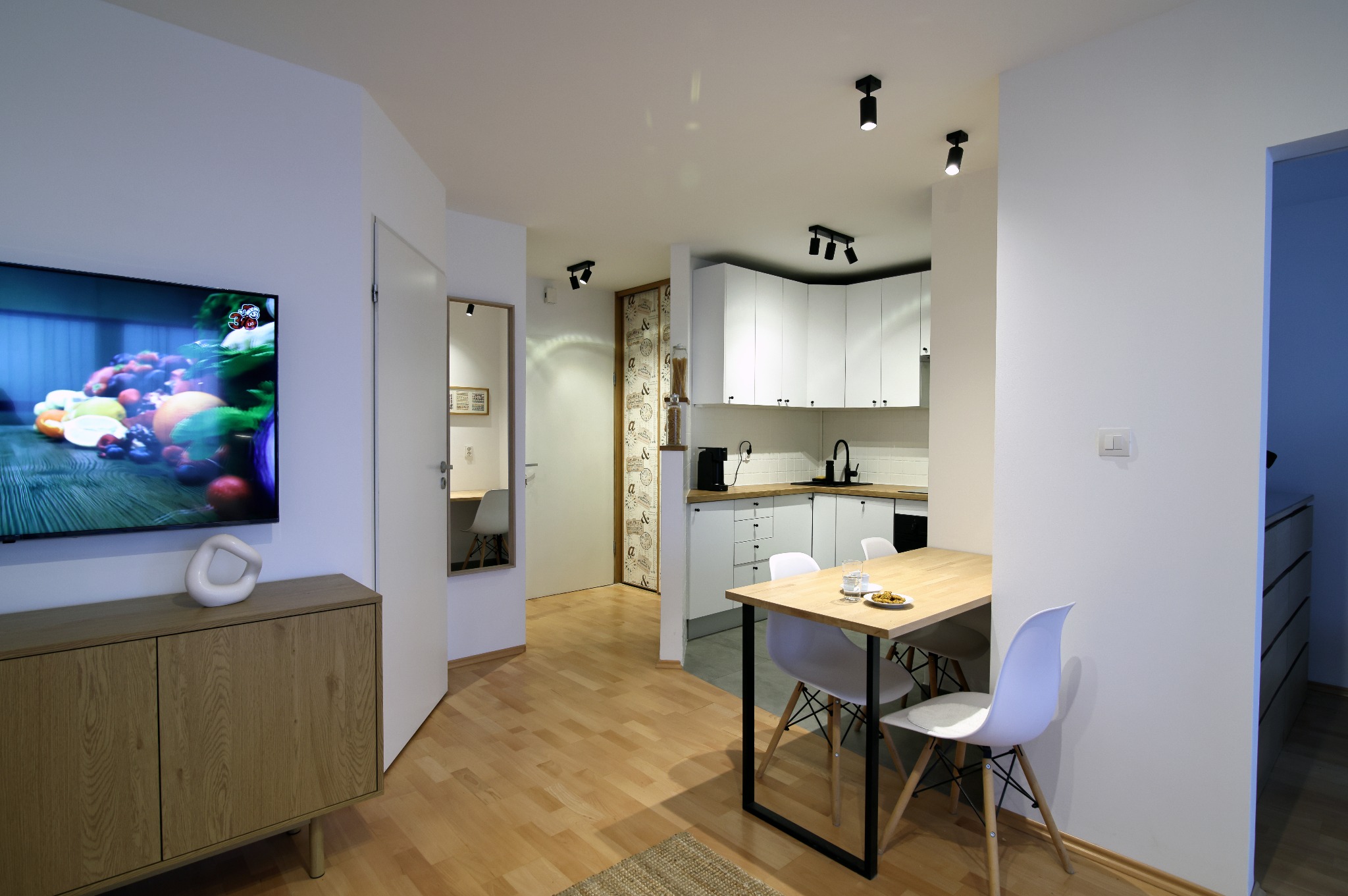 Jasne wnętrze mieszkania: drewniana podłoga, biała kuchnia z czarnymi akcentami, stół z krzesłami i telewizor z owocami na ścianie. Nowoczesny design.