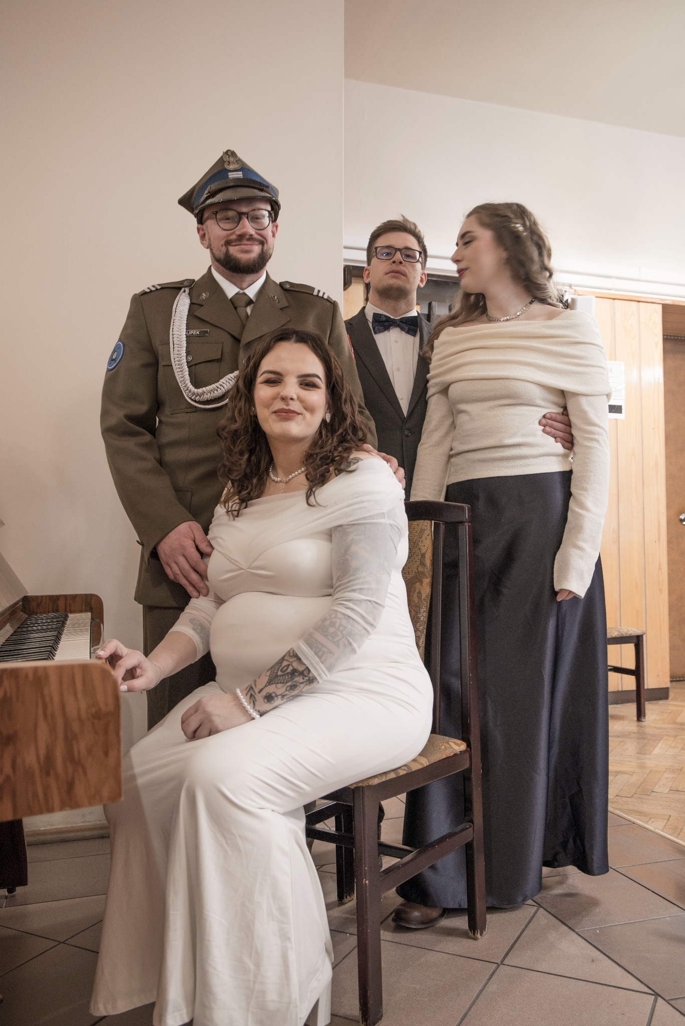 Uśmiechnięta kobieta w białej sukni siedzi przy pianinie, obok stoi mężczyzna w mundurze wojskowym, a za nimi para w strojach wieczorowych. Nietypowe ujęcie.