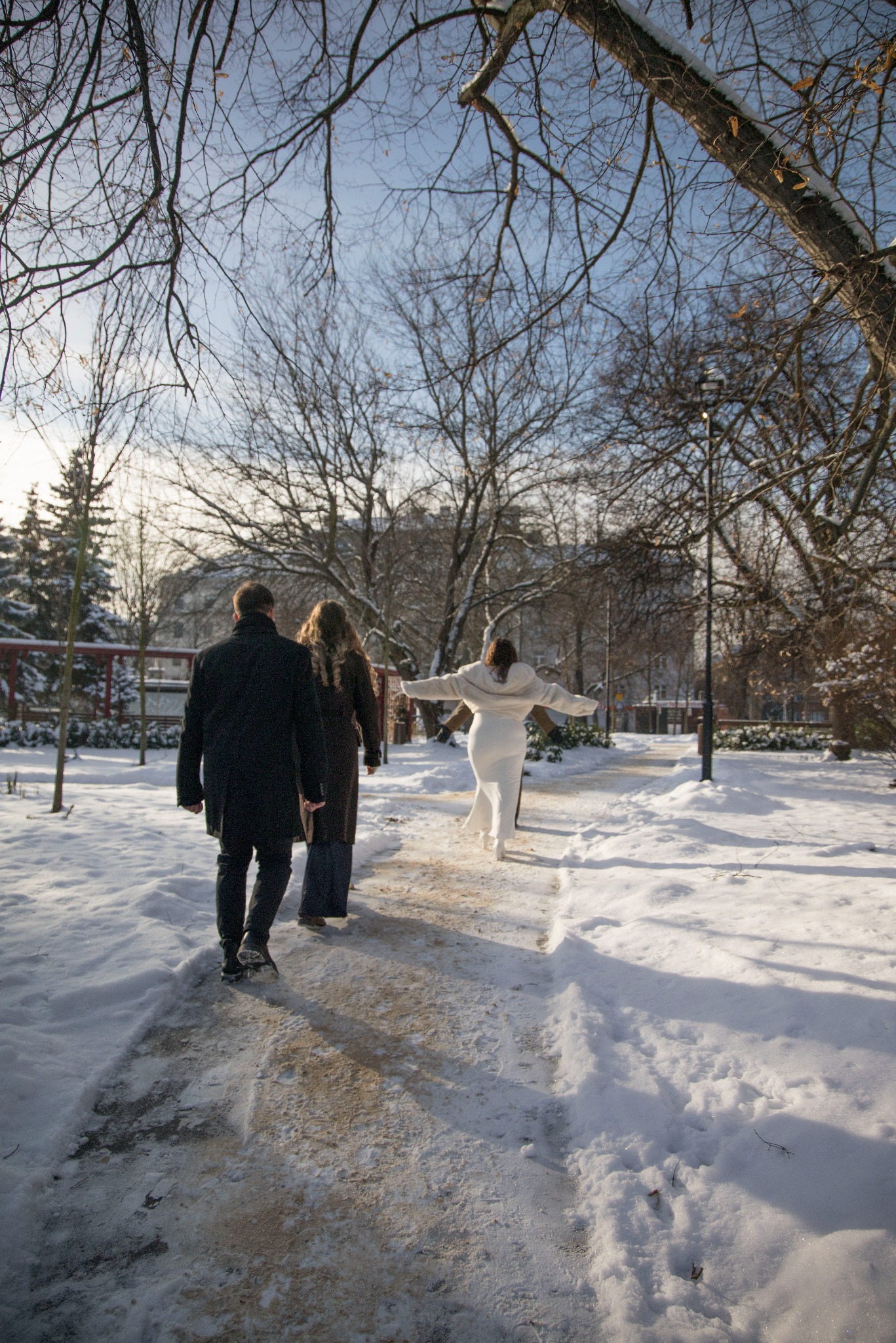 Zimowa sesja w Krakowie: Para spaceruje po ośnieżonym parku, a kobieta w białej sukience rozkłada ramiona, ciesząc się otoczeniem. Drzewa bez liści i delikatne światło słoneczne.