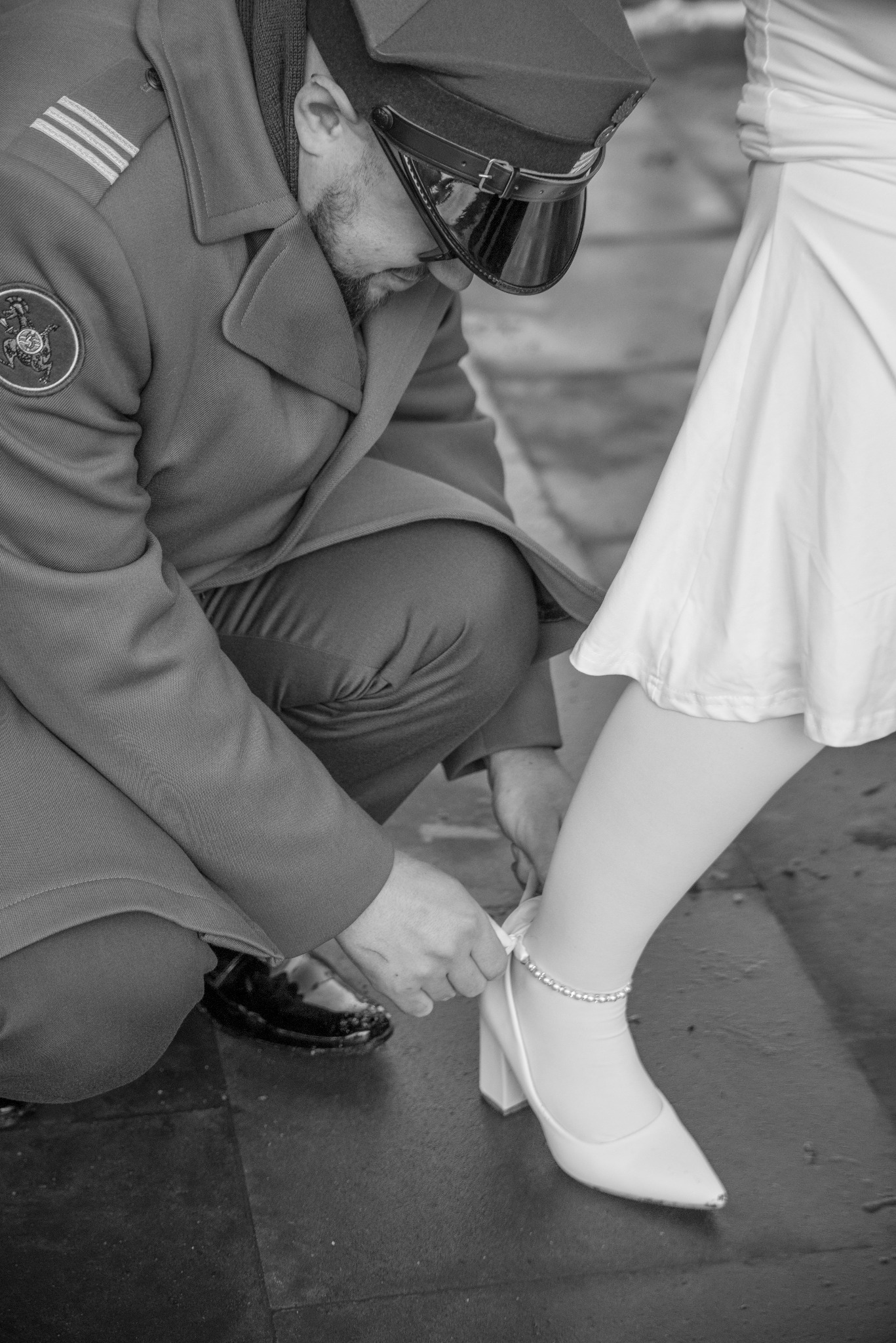 Czarno-białe zdjęcie: mężczyzna w mundurze poprawia but panny młodej z bransoletką na kostce. Ujęcie z bliska, skupione na detalu. Romantyczny moment w dniu ślubu.
