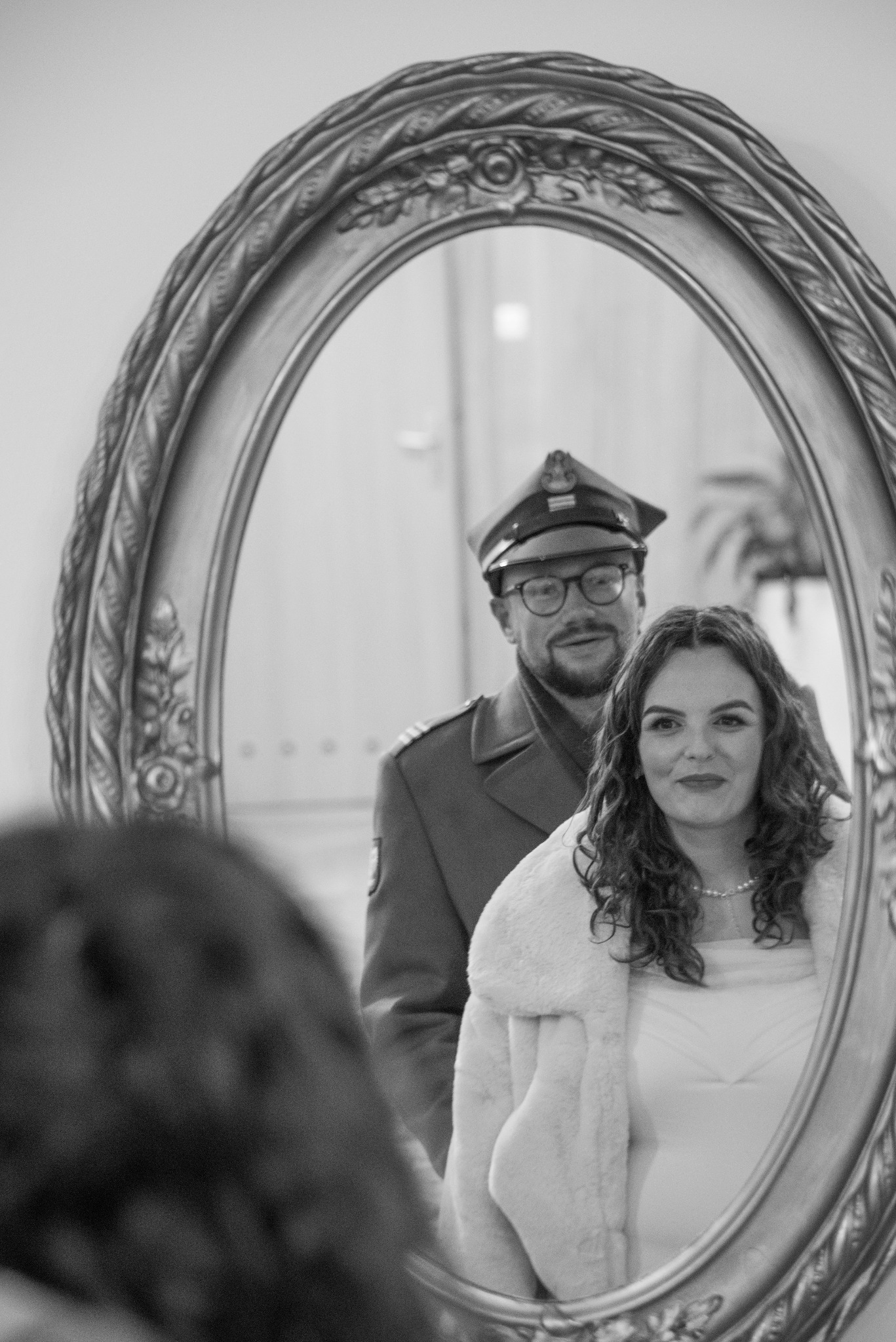 Czarno-białe odbicie w ozdobnym lustrze: uśmiechnięta panna młoda w futrze i oficer w mundurze. Nietypowe ujęcie ślubne z wojskowym akcentem.