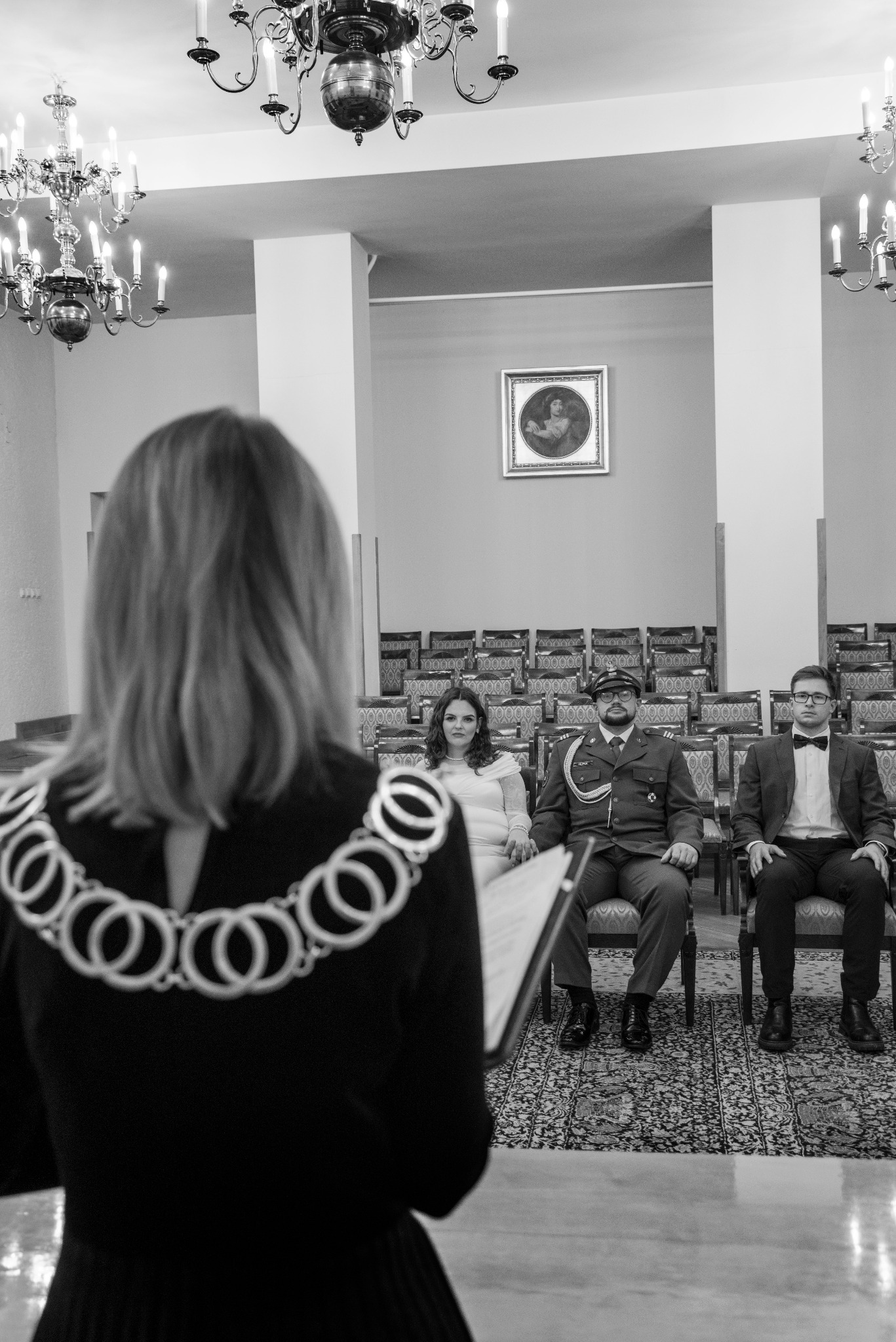 Czarno-białe zdjęcie z ceremonii ślubnej: urzędniczka z łańcuchem, para młoda i świadek siedzą na krzesłach w sali z żyrandolami i obrazem na ścianie.