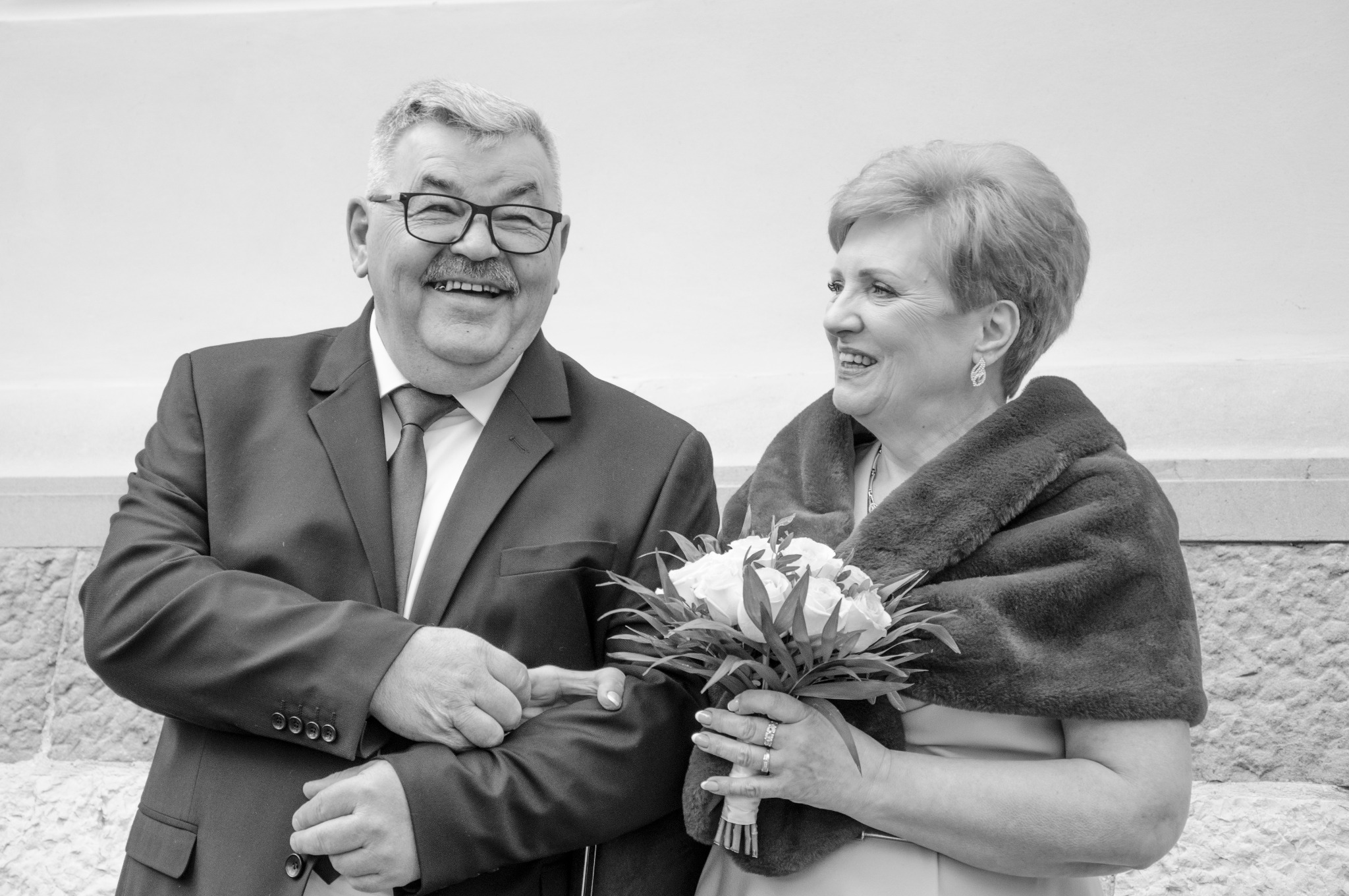 Uśmiechnięta para w strojach formalnych, trzymająca się za ręce i bukiet białych róż. Zdjęcie czarno-białe, z widocznymi detalami twarzy i fakturą ubrań.