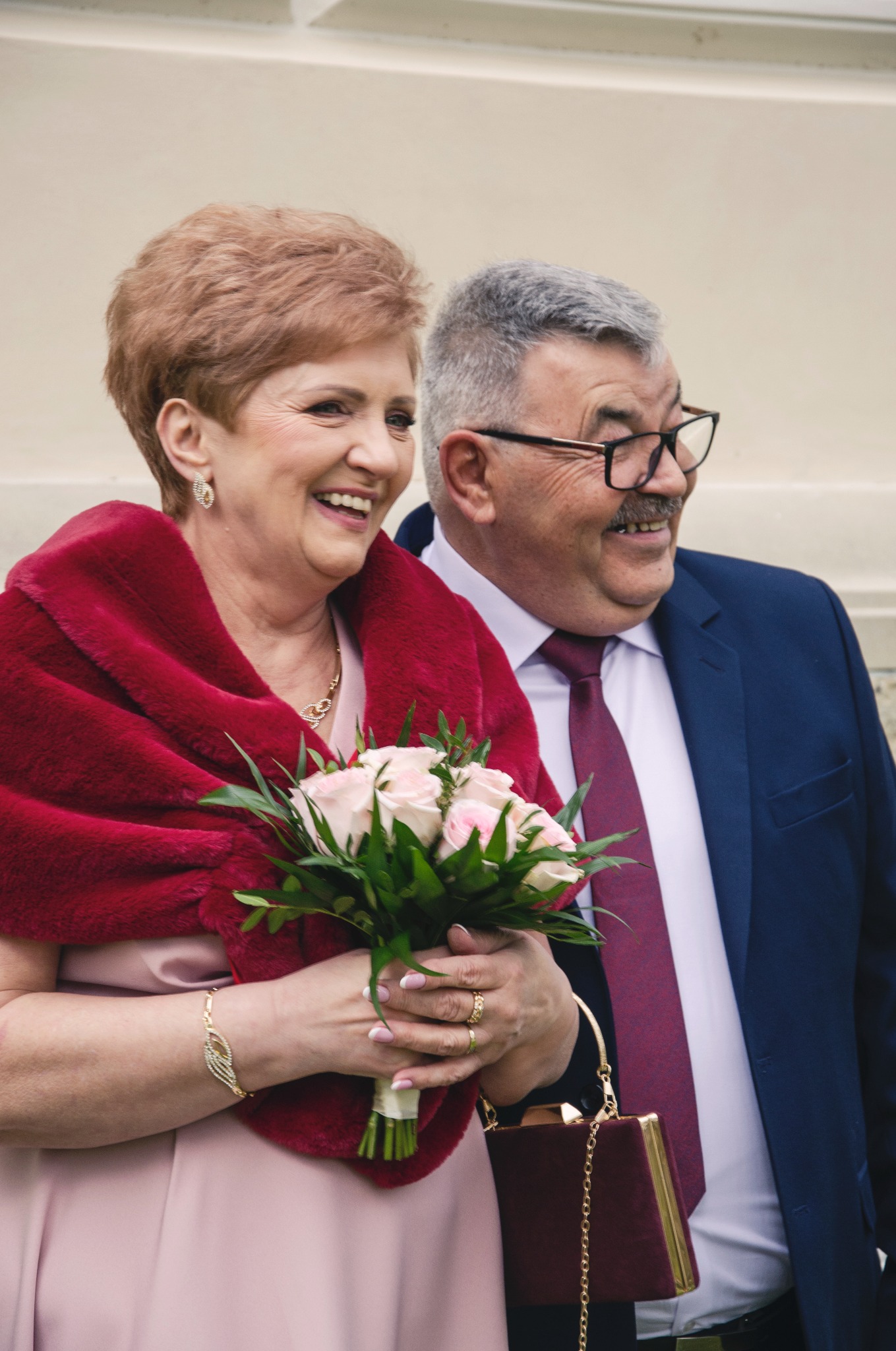 Uśmiechnięta para w eleganckich strojach: kobieta z bukietem różowych róż i szalem, mężczyzna w garniturze. Radosne spojrzenie w bok, jasne tło.