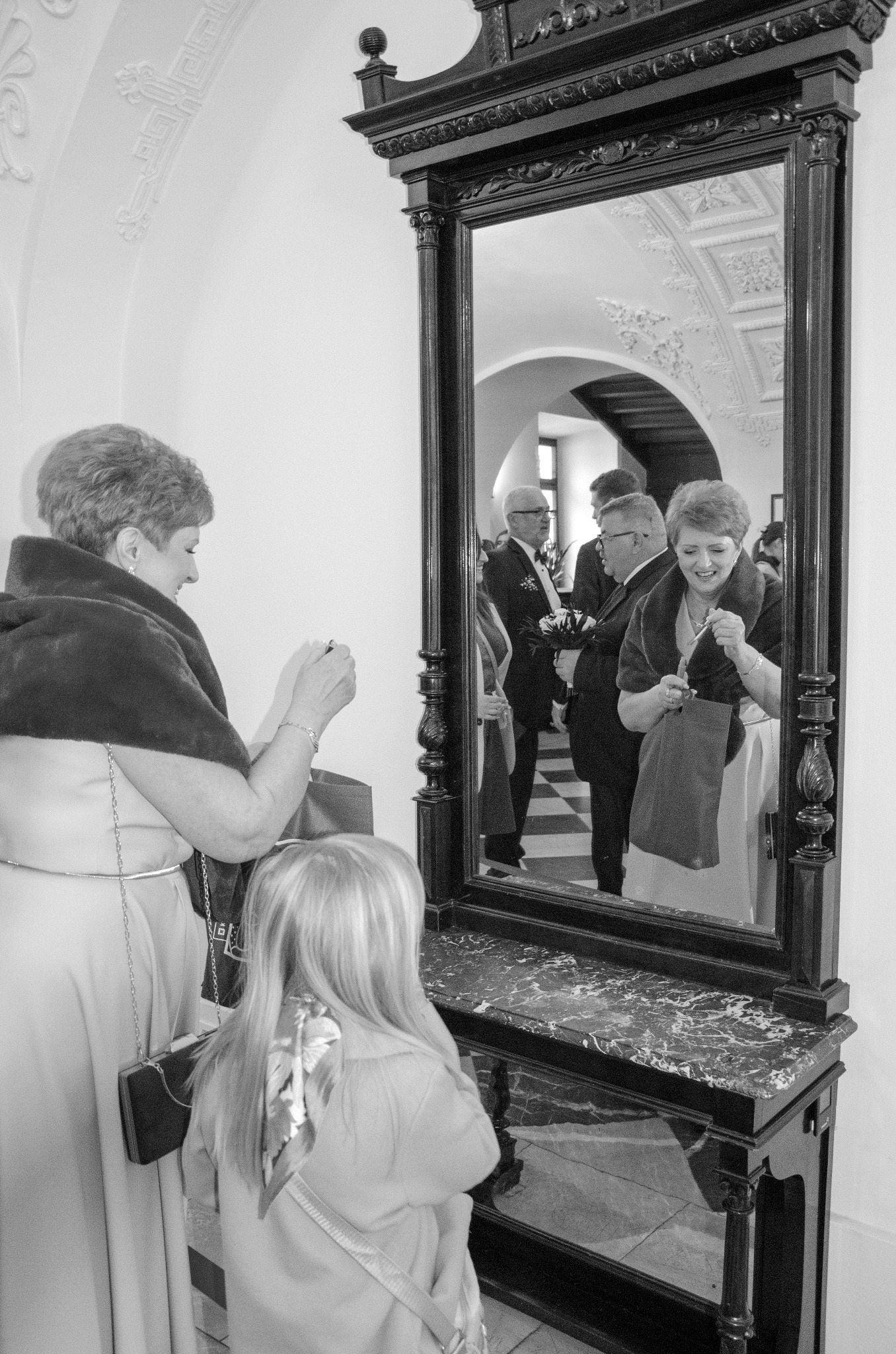 Czarno-białe zdjęcie kobiety robiącej zdjęcie w lustrze, w tle widoczni goście w eleganckich strojach. Dziewczynka patrzy na lustro. Marmurowy blat konsoli.